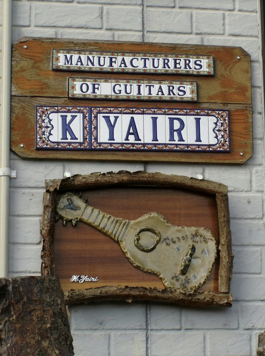 Signboard reading 'Manufacturers of Guitars' with decorative tiles spelling 'KYAIRI' and a wooden cutout of a guitar on a stone wall.