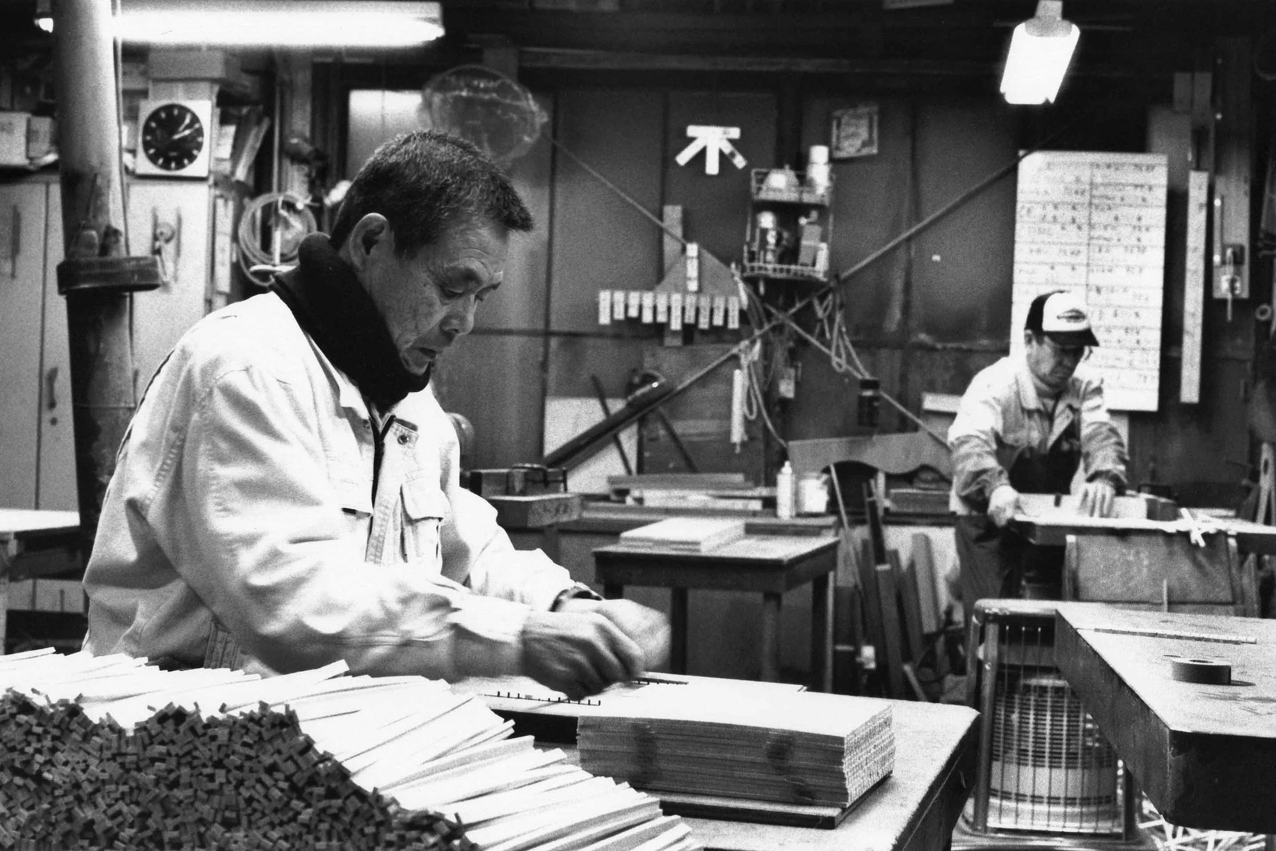 Two men working in a woodworking workshop, sorting and organizing wood pieces, with tools and equipment hanging on the wall.
