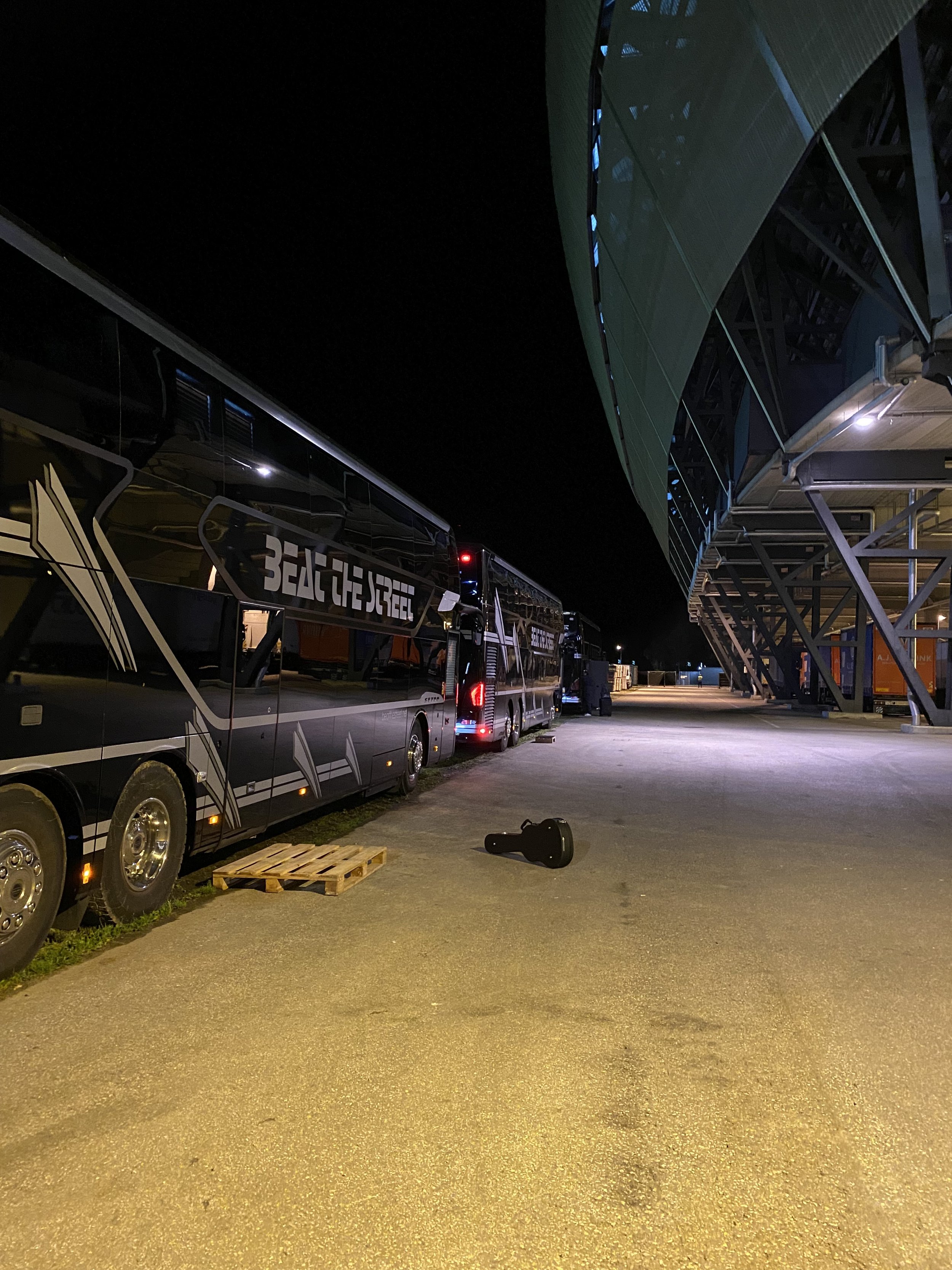 Nighttime scene featuring a large bus with 'BEAT THE STEEL' on its side parked beside a modern building with a unique green facade and large glass windows. Grace waiting on the ground nearby, with construction or storage containers in the background.