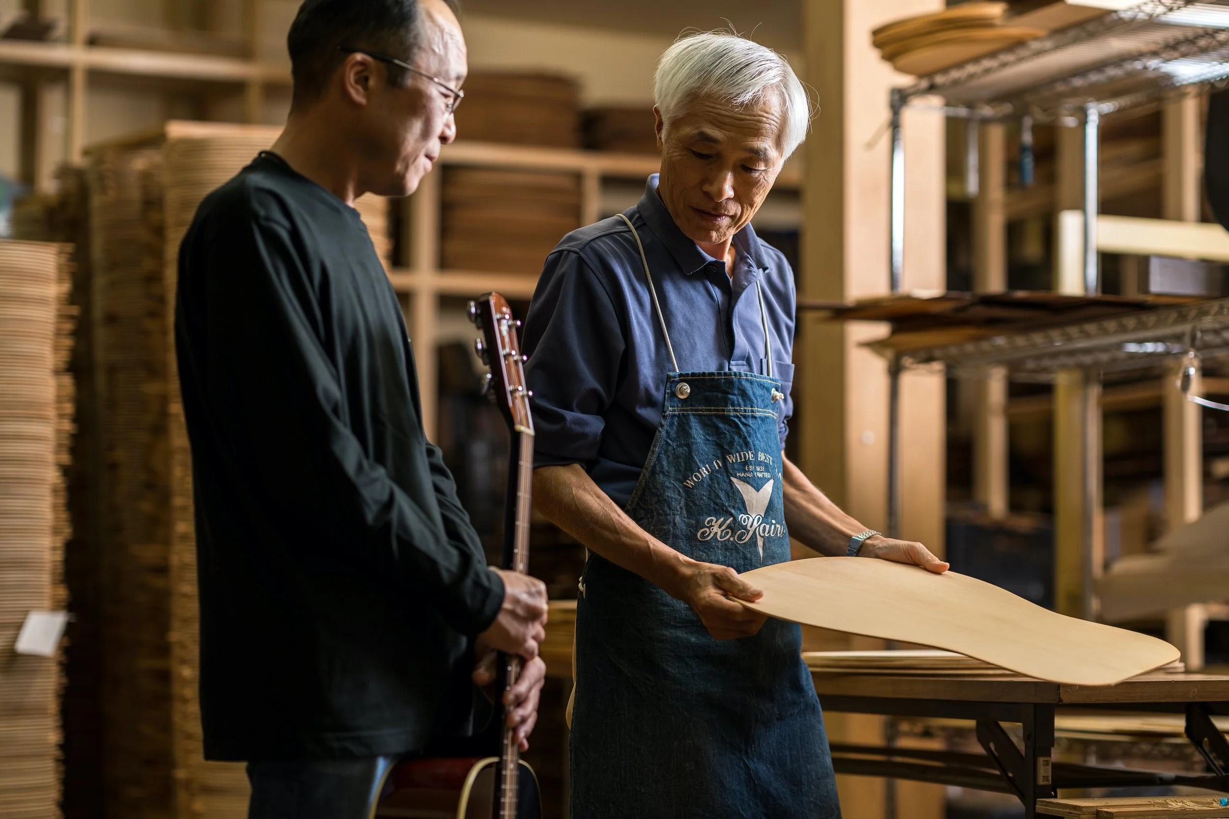 Two men in a woodworking shop examining a large piece of wood, with shelves of wood panels in the background.