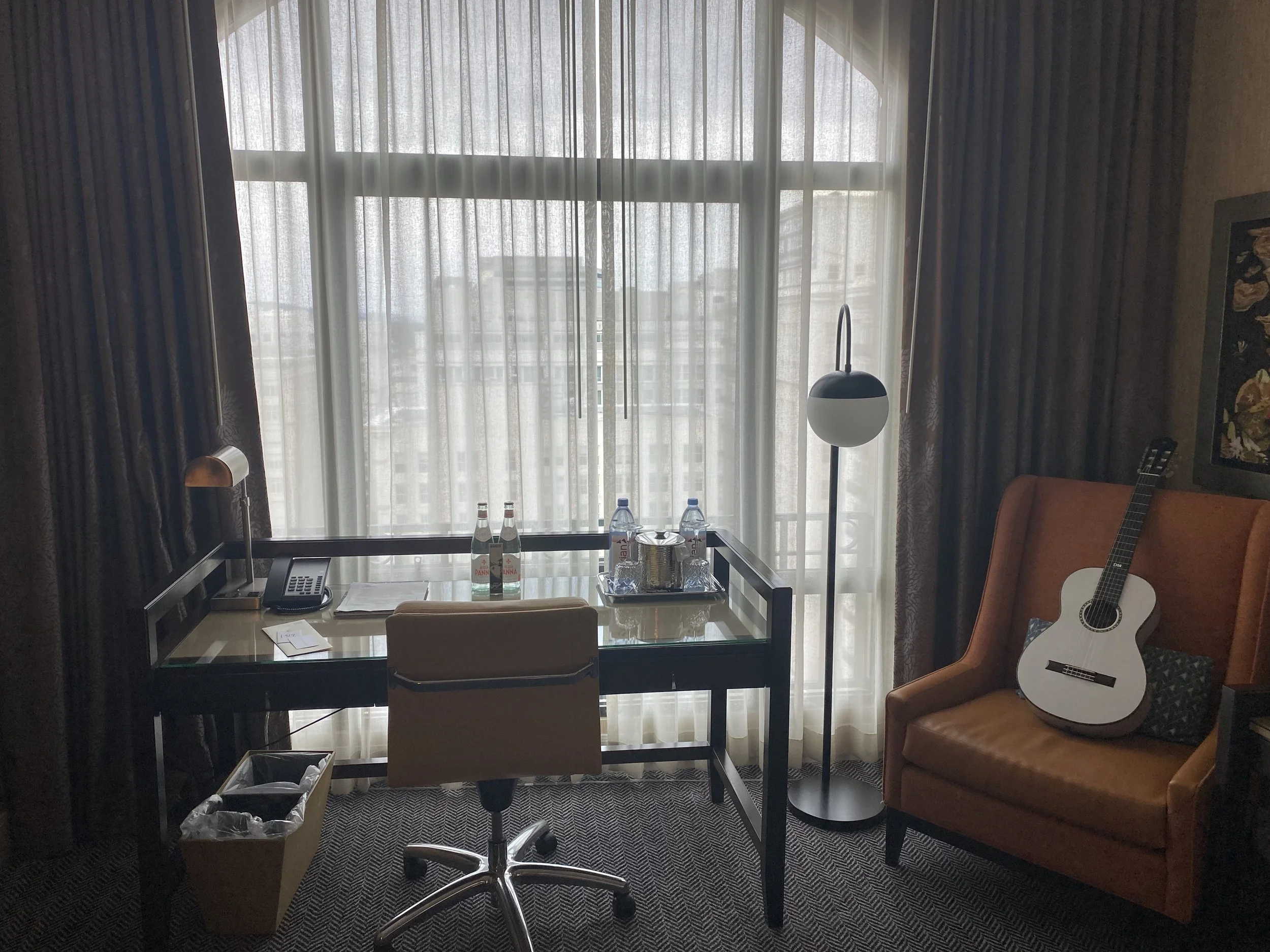 A hotel with a large window covered by sheer curtains, a desk with water bottles and a phone, a tan leather chair with Grace, a floor lamp, and a framed artwork on the wall.