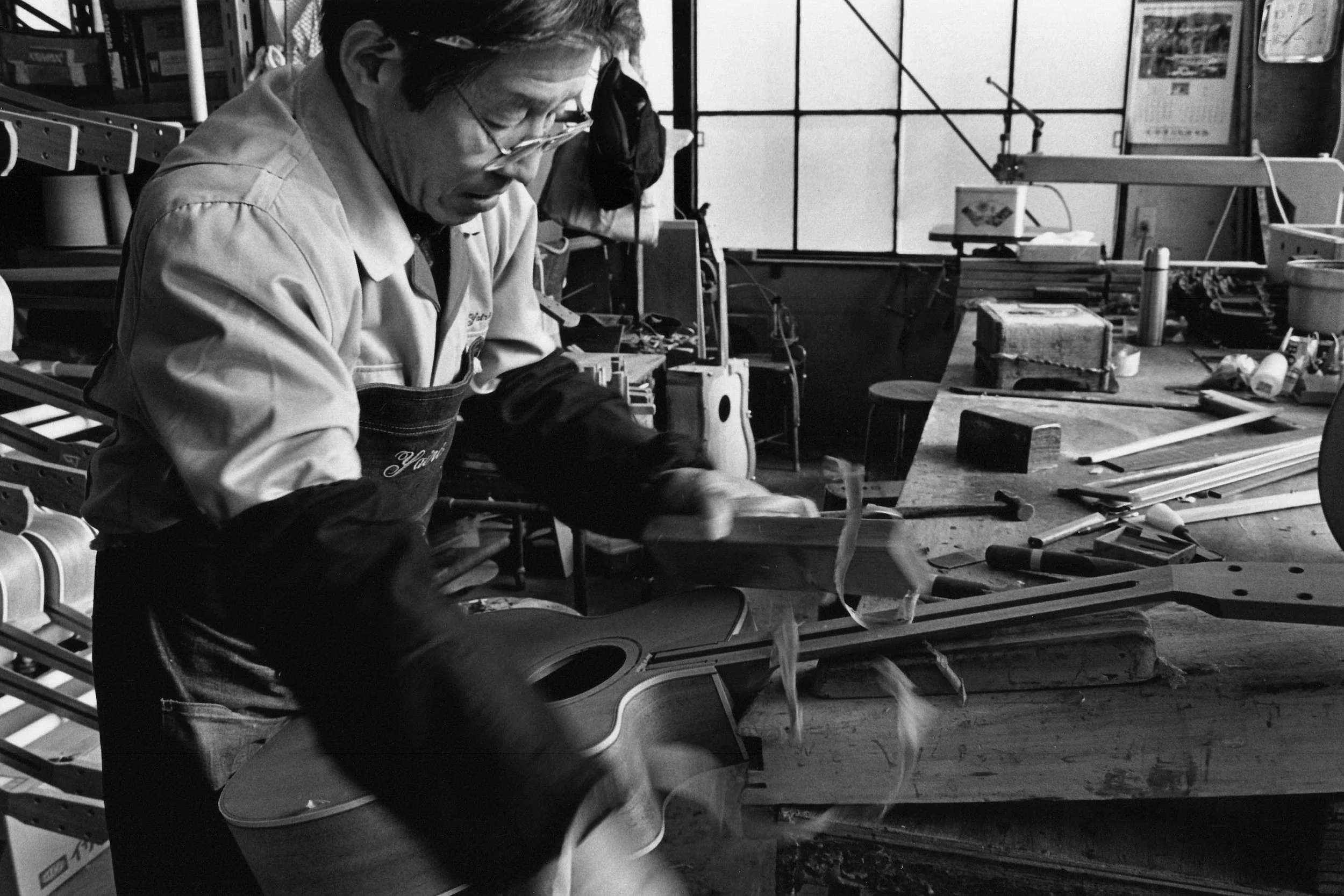 A man working in a workshop, wearing glasses and an apron, surrounded by tools and materials, with shelves and large windows in the background.