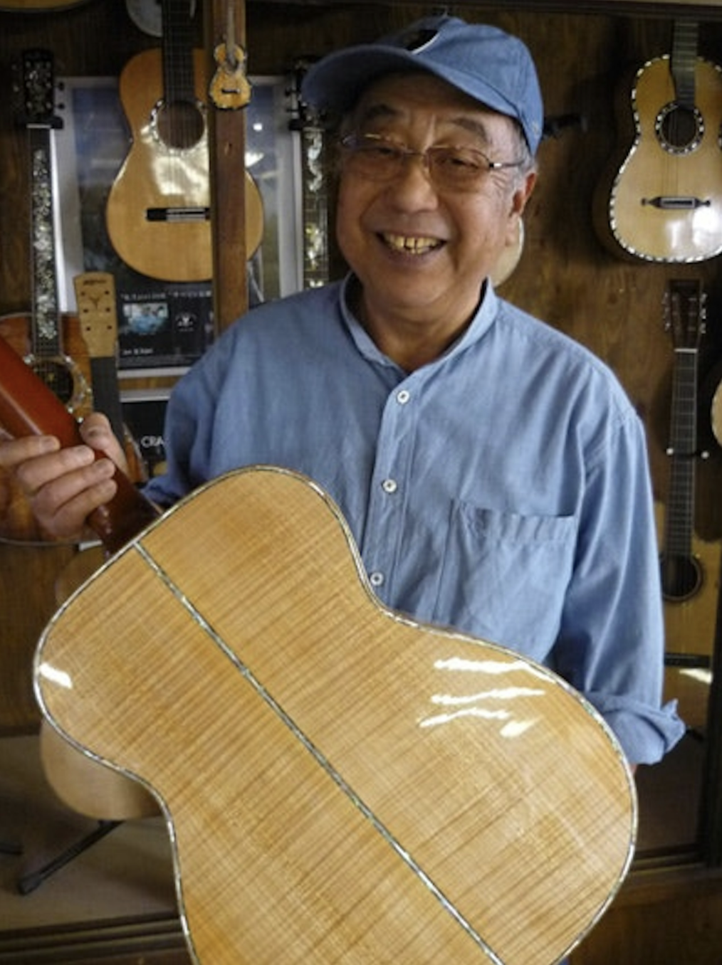 An older man smiling, wearing a blue baseball cap and glasses, holding an acoustic guitar in a music store with other guitars hanging on the wall behind him.
