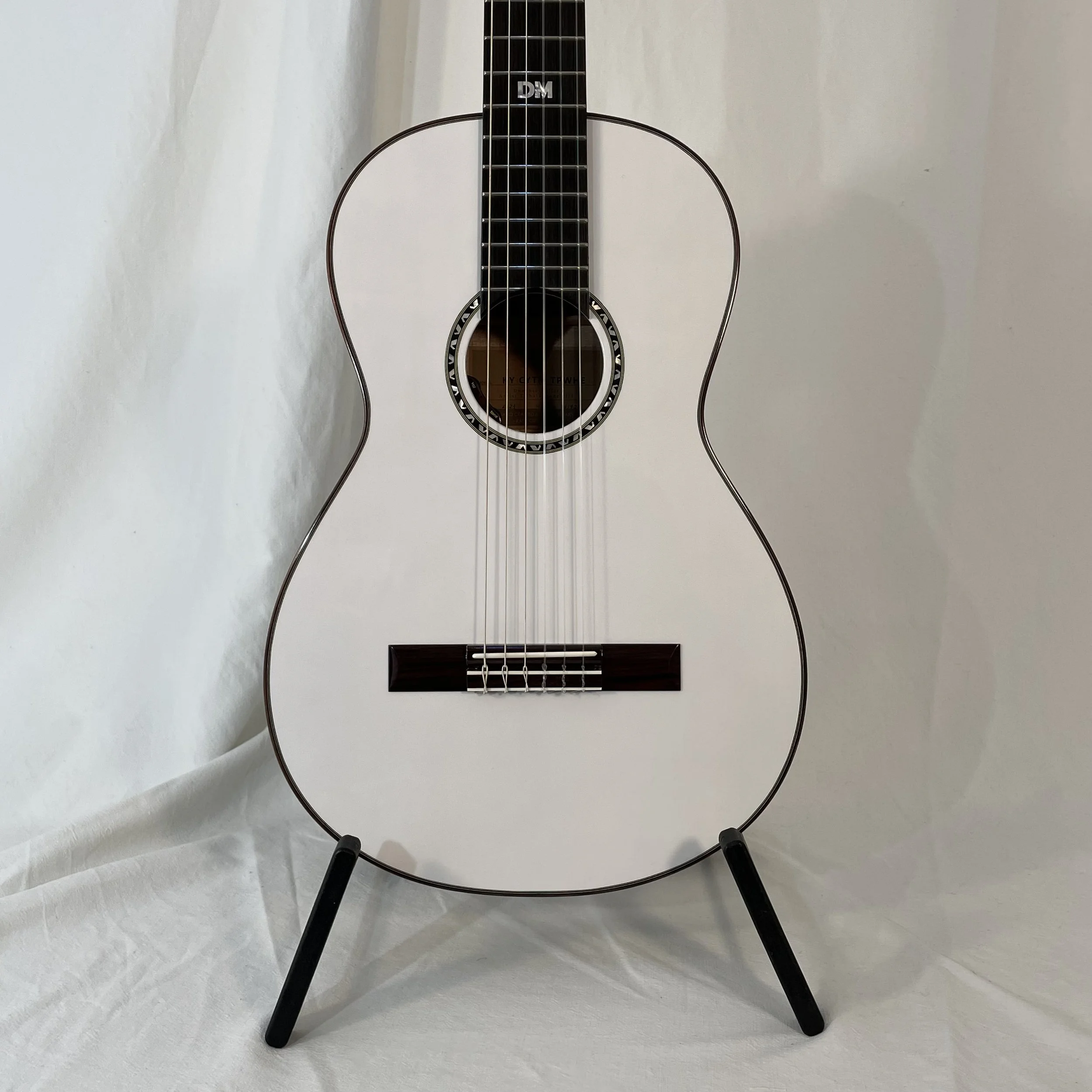 An acoustic guitar with a light-colored body, dark fretboard, and a soundhole with decorative inlay, resting on a black, metal, circular stand with three legs, placed on a white fabric surface against a plain white background.
