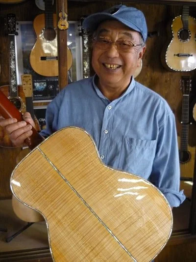 Kazuo Yairi smiling, wearing a blue baseball cap and glasses, holding an acoustic guitar in a music store with other guitars hanging on the wall behind him.