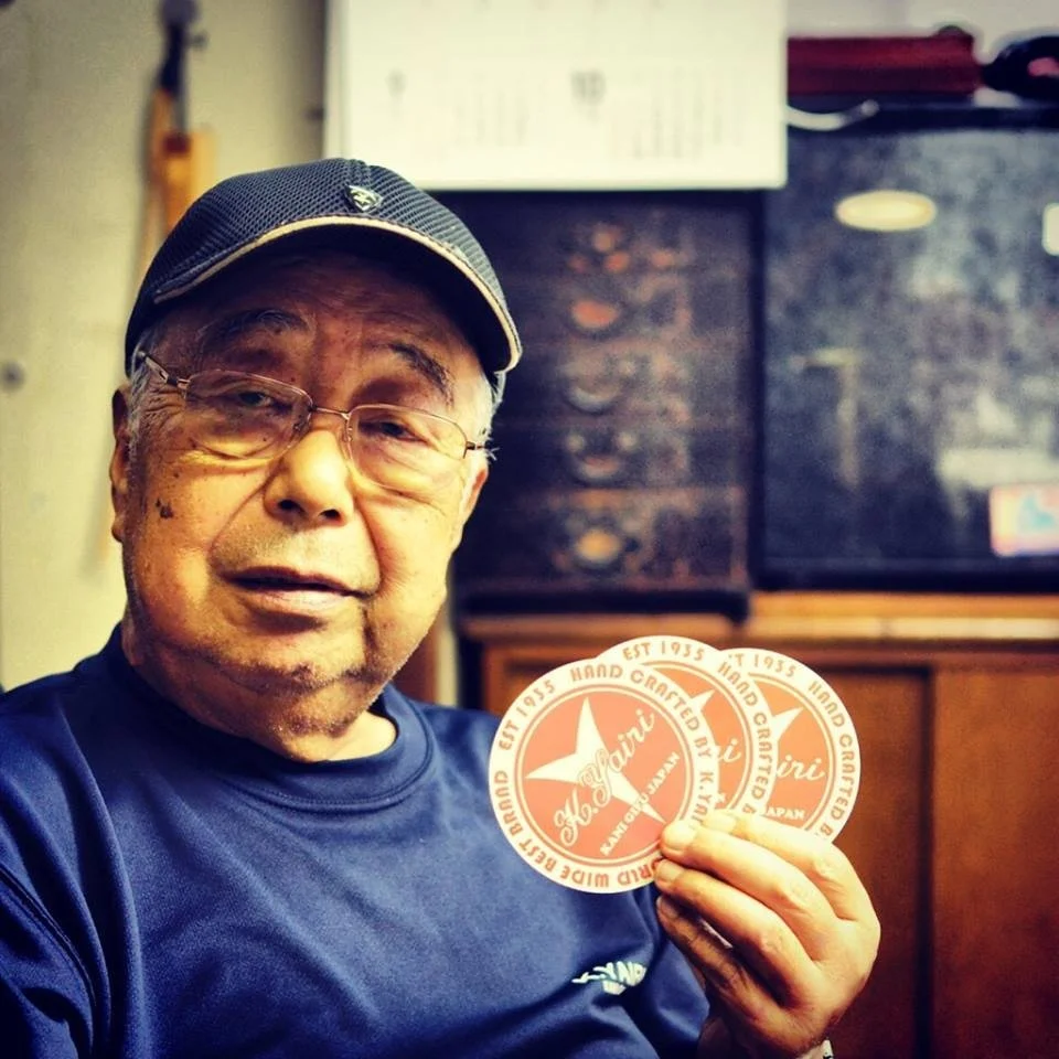 An older man with glasses and a black cap holding up a round sticker with red and white colors and Japanese text, inside a room with wooden furniture and black speakers.