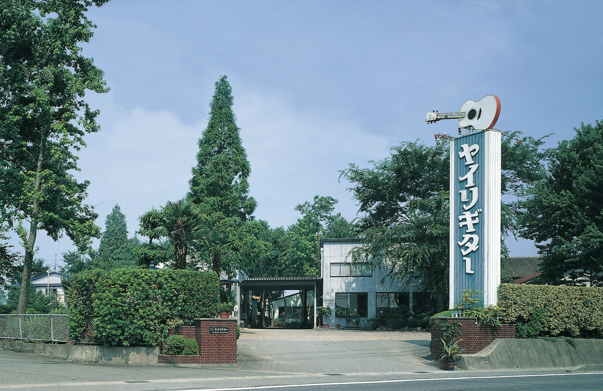 A street view with lush green trees and a building in the background. There is a tall sign with Japanese characters and a large guitar on top, indicating a music-related business. The sky is blue with some clouds.