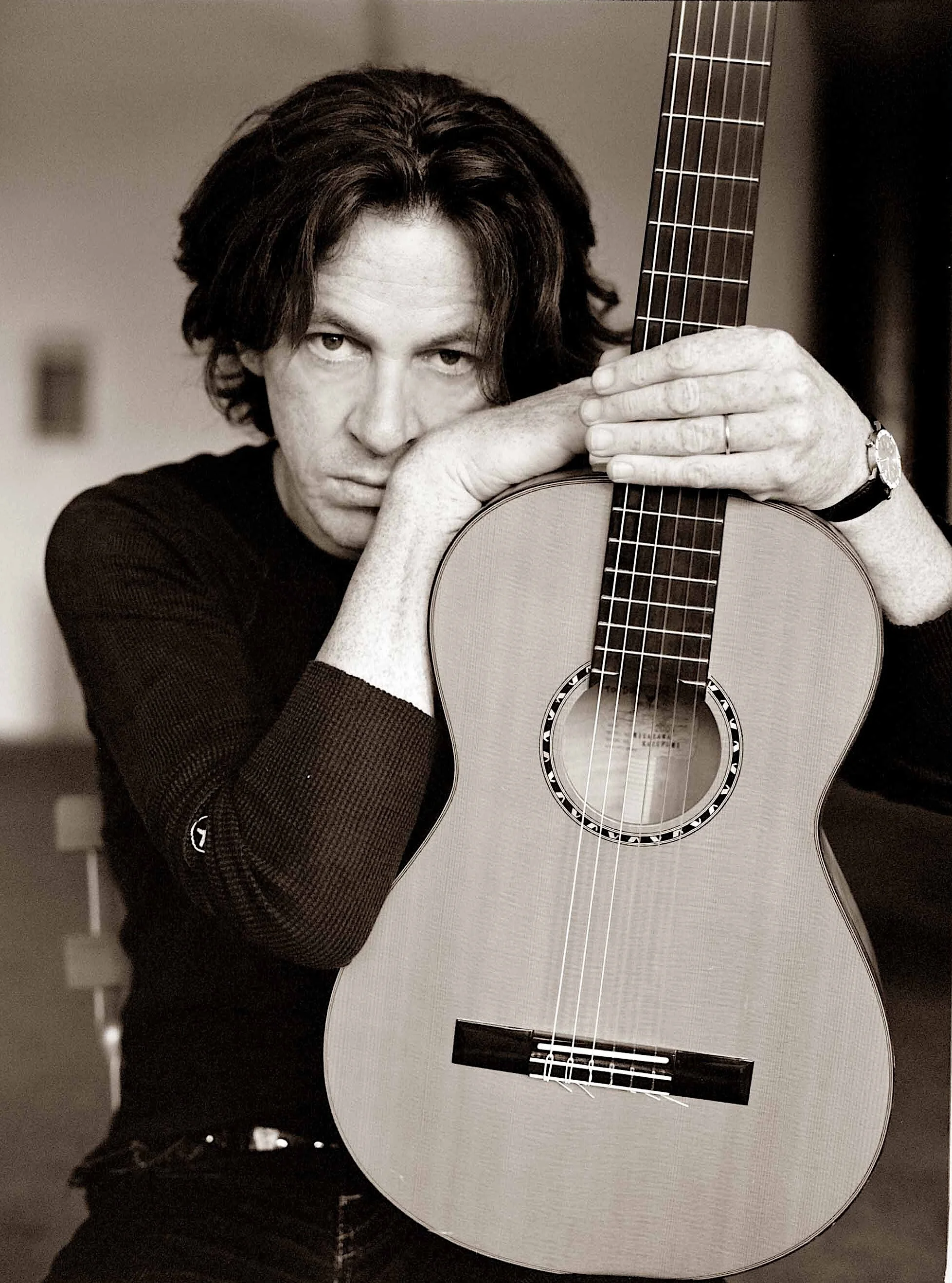 Black-and-white photo of a man with medium-length hair, holding an acoustic guitar close to his face. His eyes are visible over the guitar, and he appears to be looking directly at the camera.