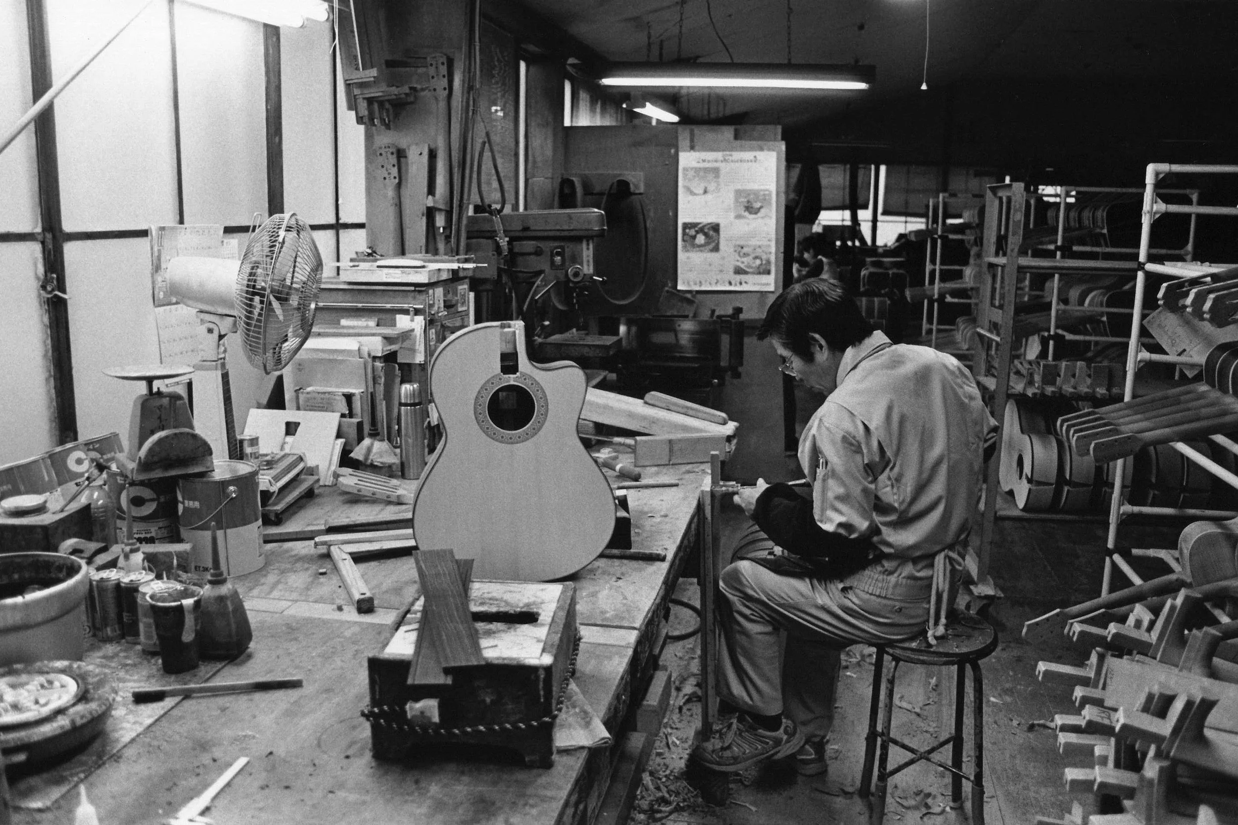 A man is working in a woodworking shop, seated at a workbench with an acoustic guitar he is building or repairing, surrounded by various tools, supplies, and materials.