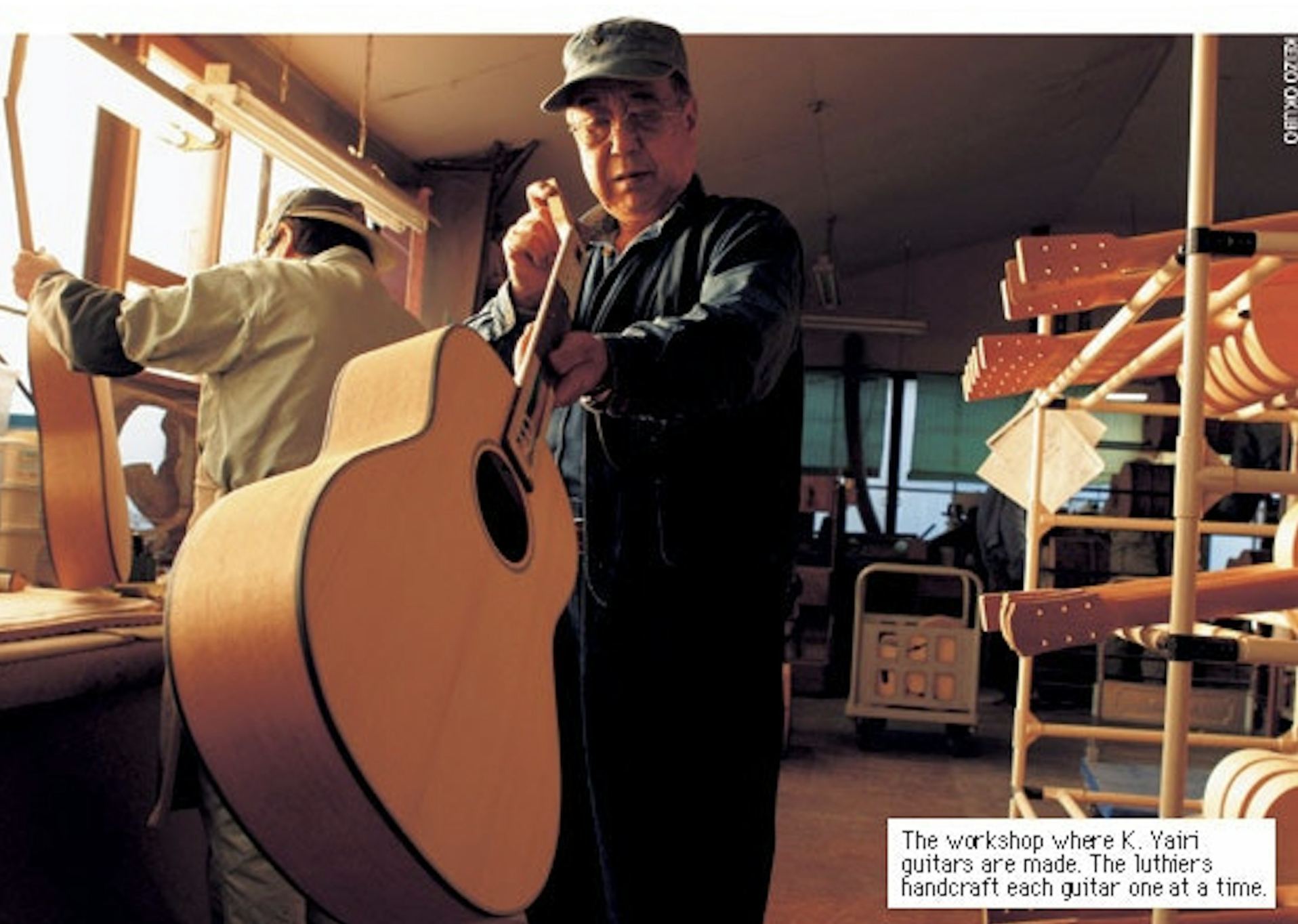A man holding a guitar in a workshop. The workshop has wooden chairs stacked on the right side and large windows on the left. The man is wearing glasses, a black jacket, and a cap, and is looking at the camera.
