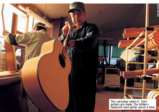 Kazuo Yairi smiling, wearing a blue baseball cap and glasses, holding an acoustic guitar in a music store with other guitars hanging on the wall behind him.