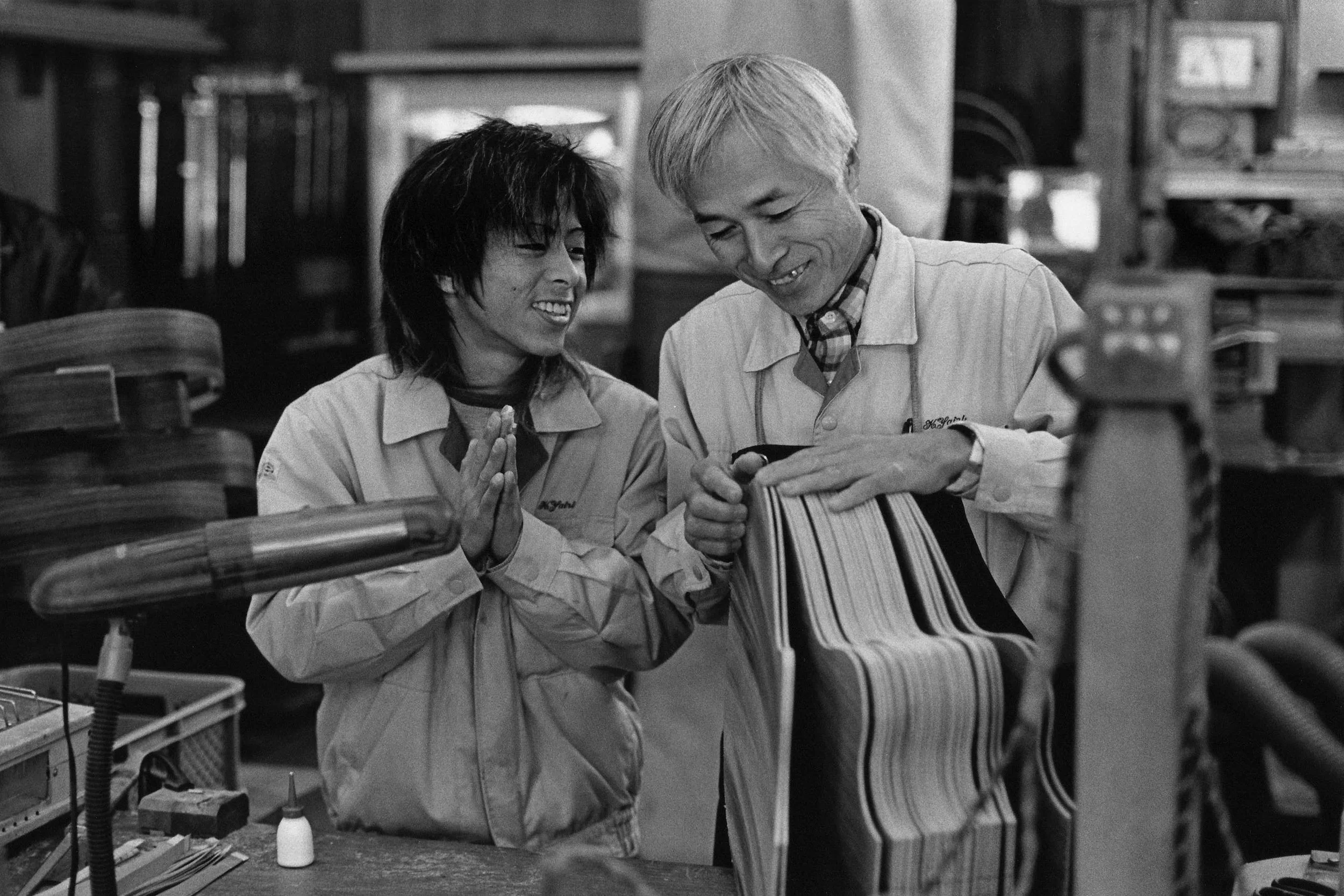 Black and white photo of two people in work uniforms leaning over a desk in a workshop, looking at a book together and smiling.