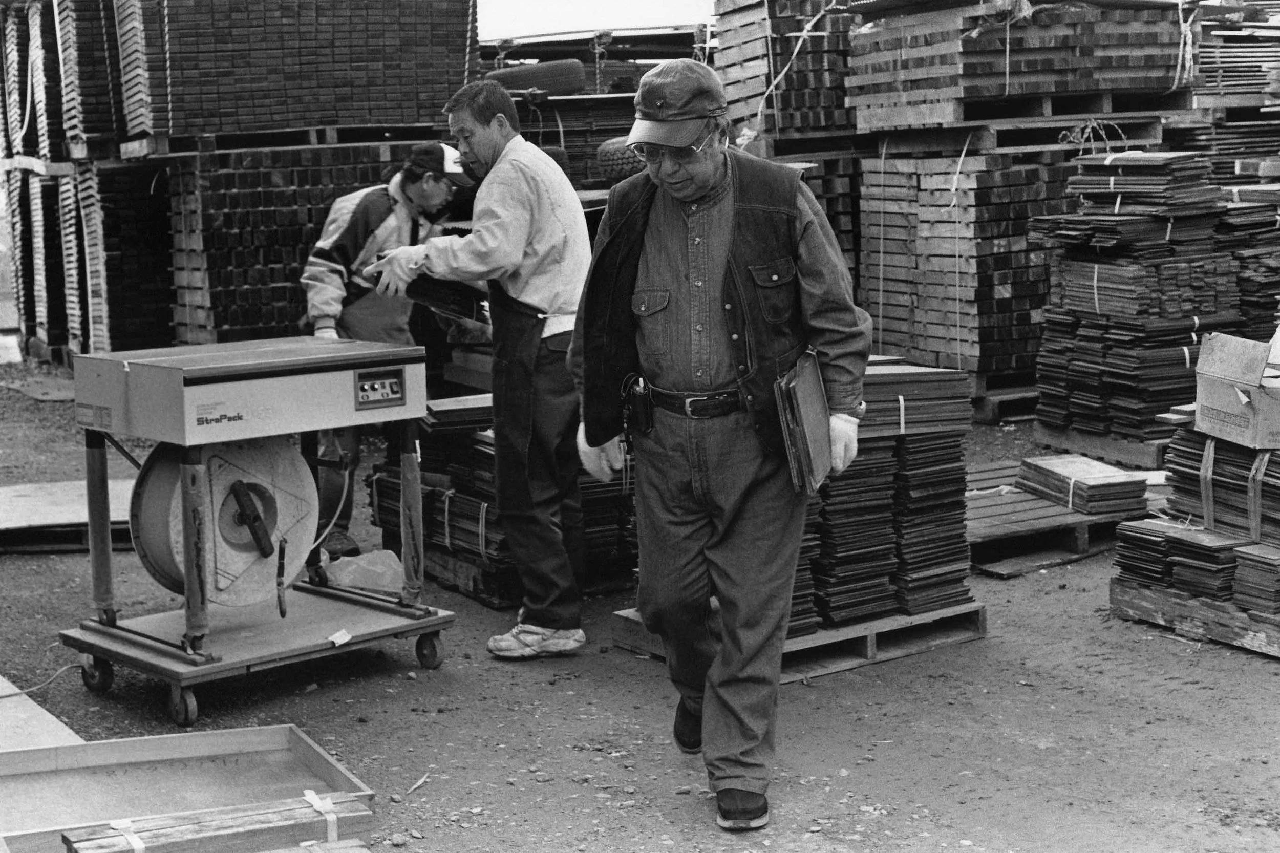 Black and white photo of three men in a lumber yard. One man is walking in the foreground holding a clipboard, while two others are looking at a device on a table. Stacks of wood and lumber are visible in the background.
