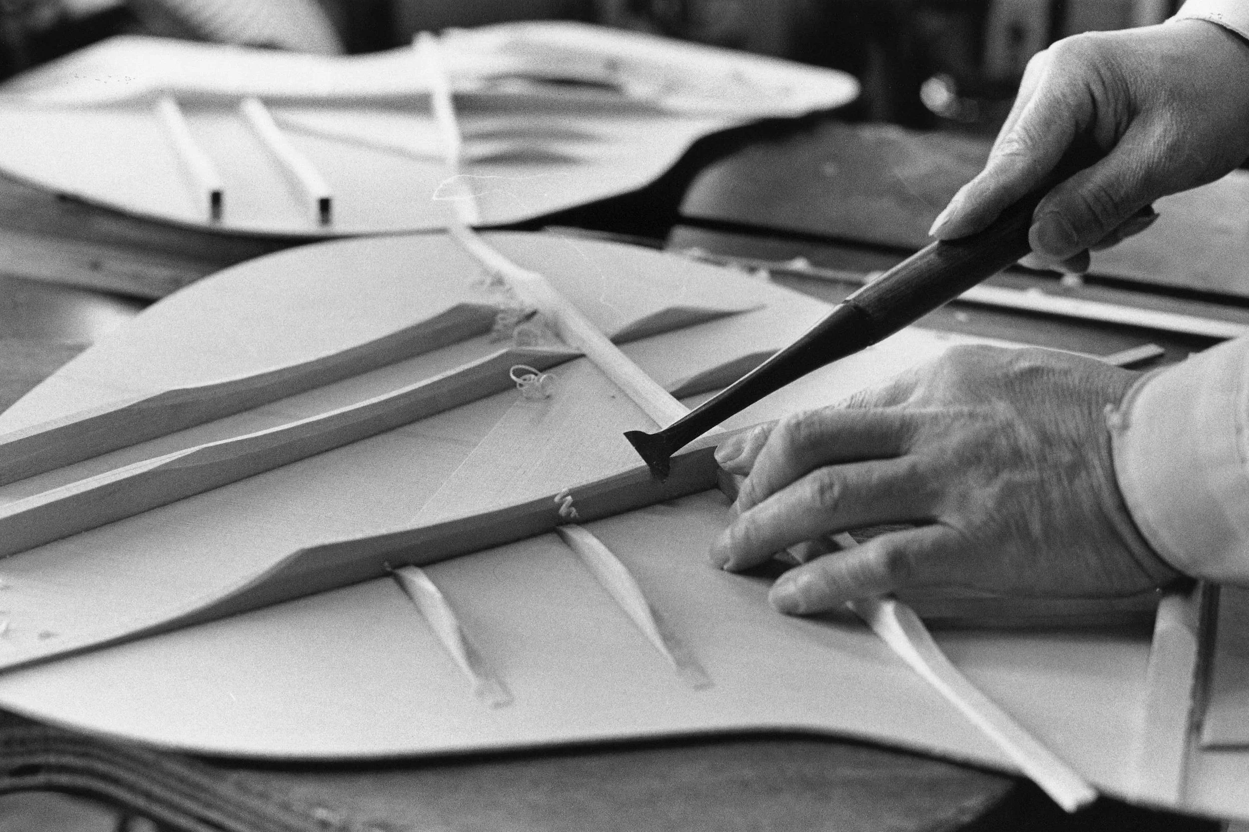 A person's hand holding a cutting tool over a stack of papers or cardboard with cut-out shapes, possibly in a workshop or design environment.