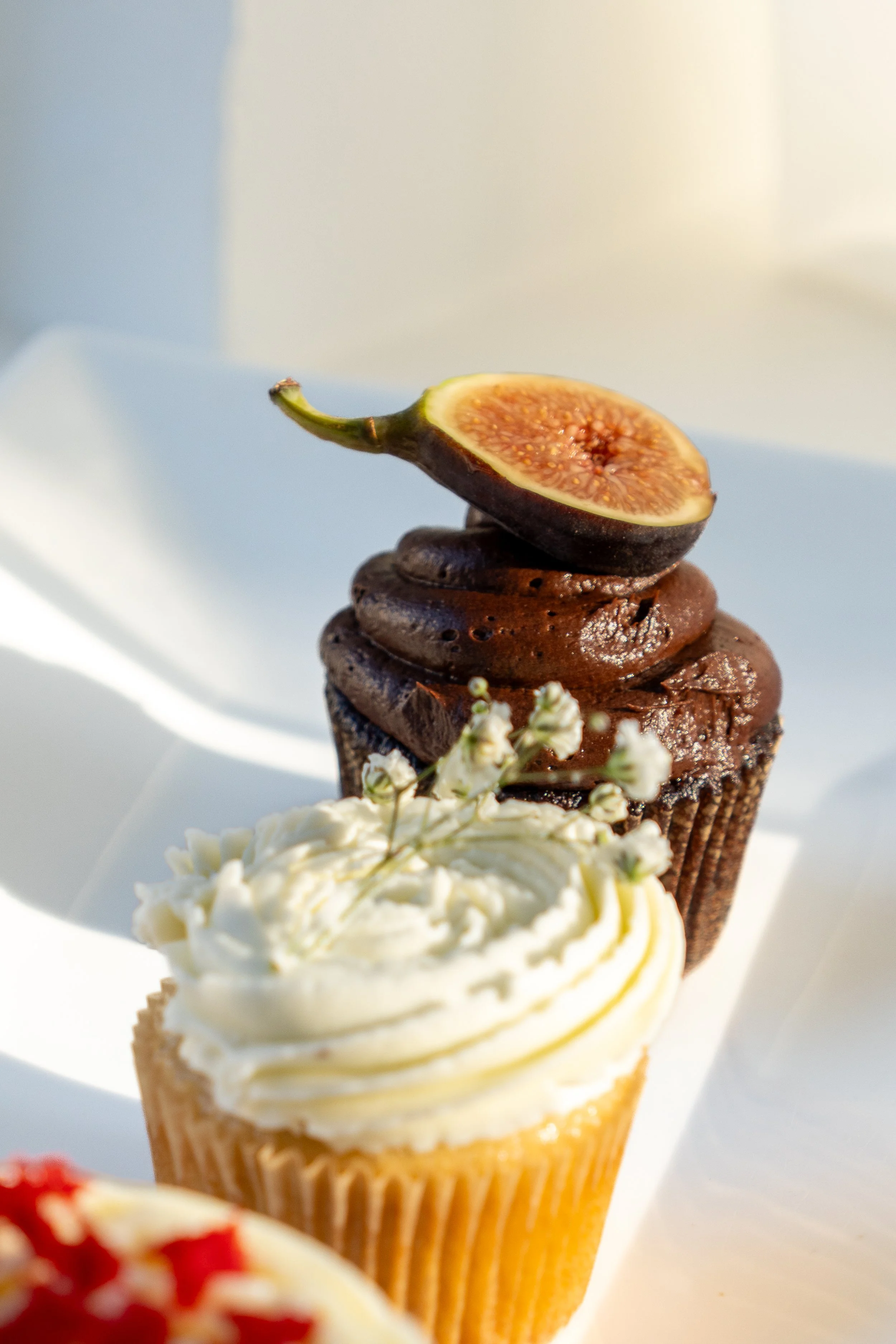 Close-up of a cupcake with vanilla frosting, topped with a halved fig, and small white flowers nearby, against a blurred light background.