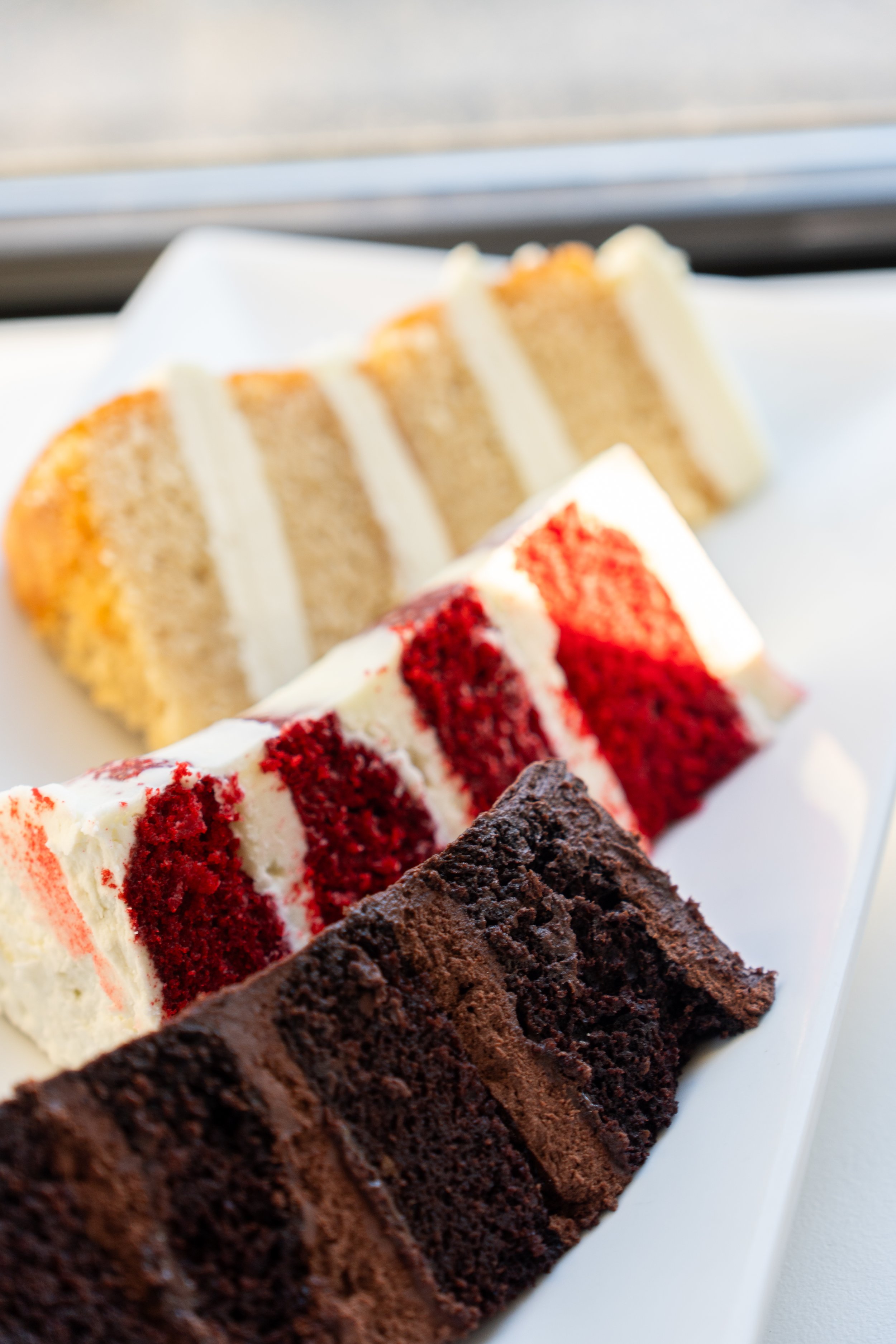 Close-up of assorted slices of cake on a white plate, including chocolate, red velvet, vanilla, and possibly carrot cake with cream cheese frosting.