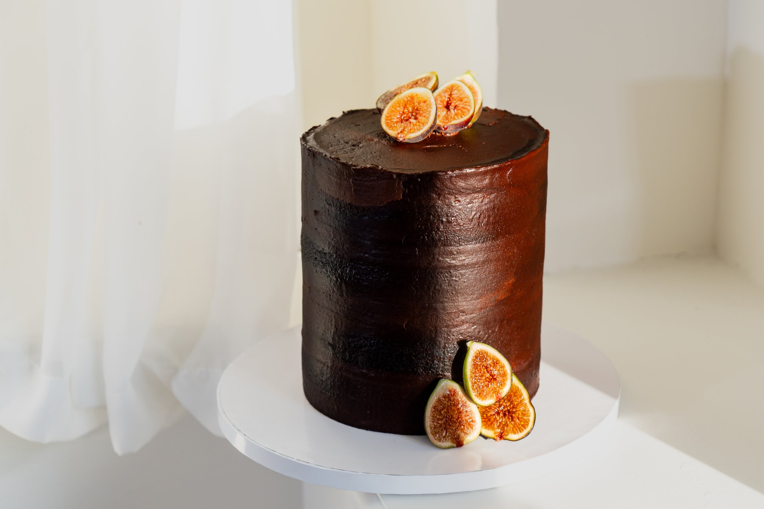 A tall, dark chocolate cake decorated with fresh fig slices on top and around the base, set on a white cake stand against a light background.