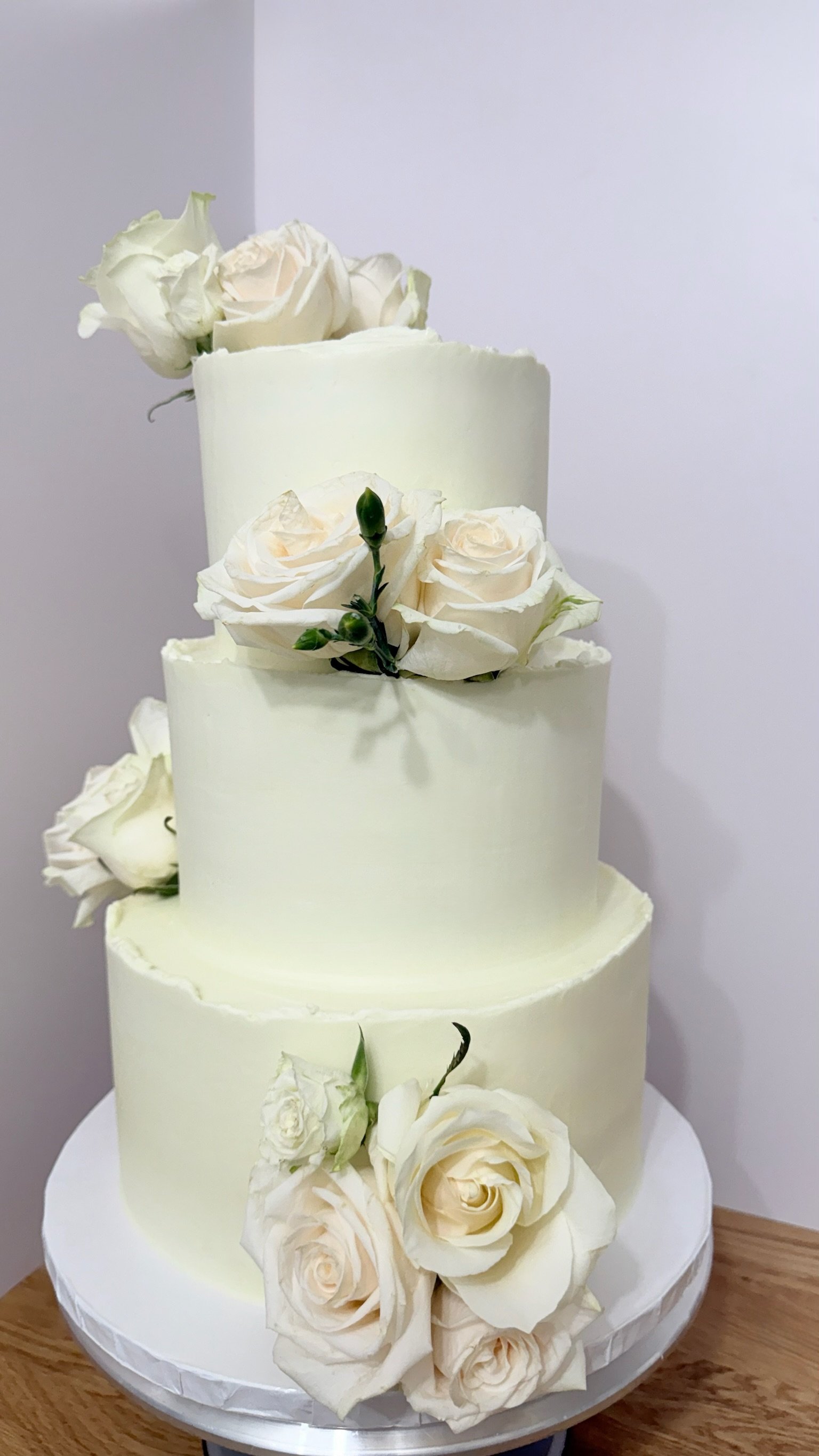 A multi-tier white wedding cake decorated with white and pale pink roses and green buds, placed on a white cake stand on a wooden surface.