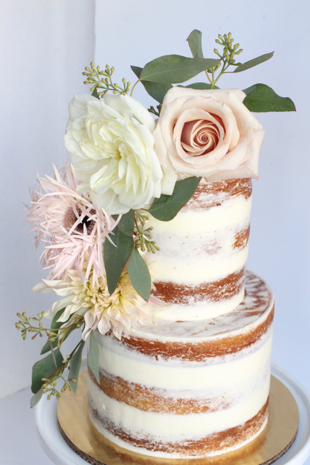 A partially frosted, multi-layer naked cake decorated with fresh flowers and greenery, with visible cake layers through the thin layer of frosting.