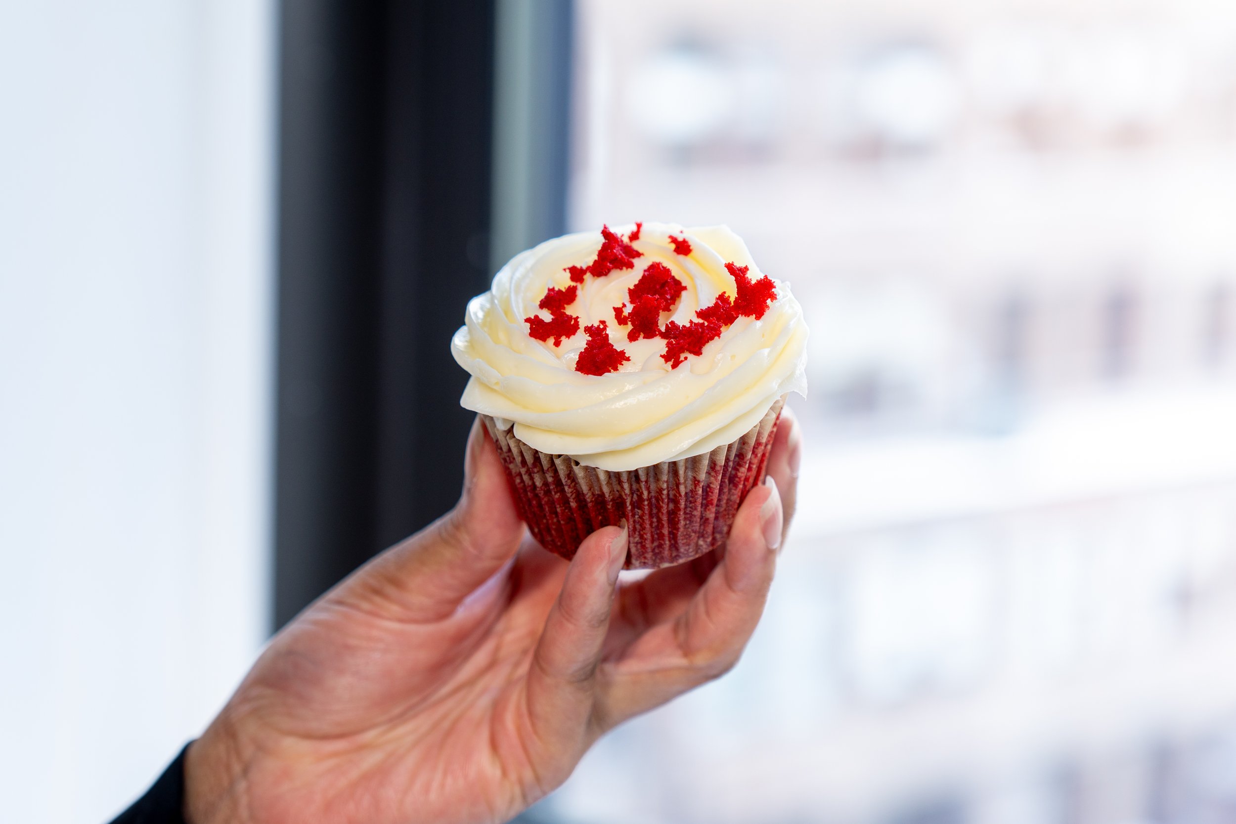 A hand holding a cupcake with white frosting and red sprinkles, with a blurred window background.