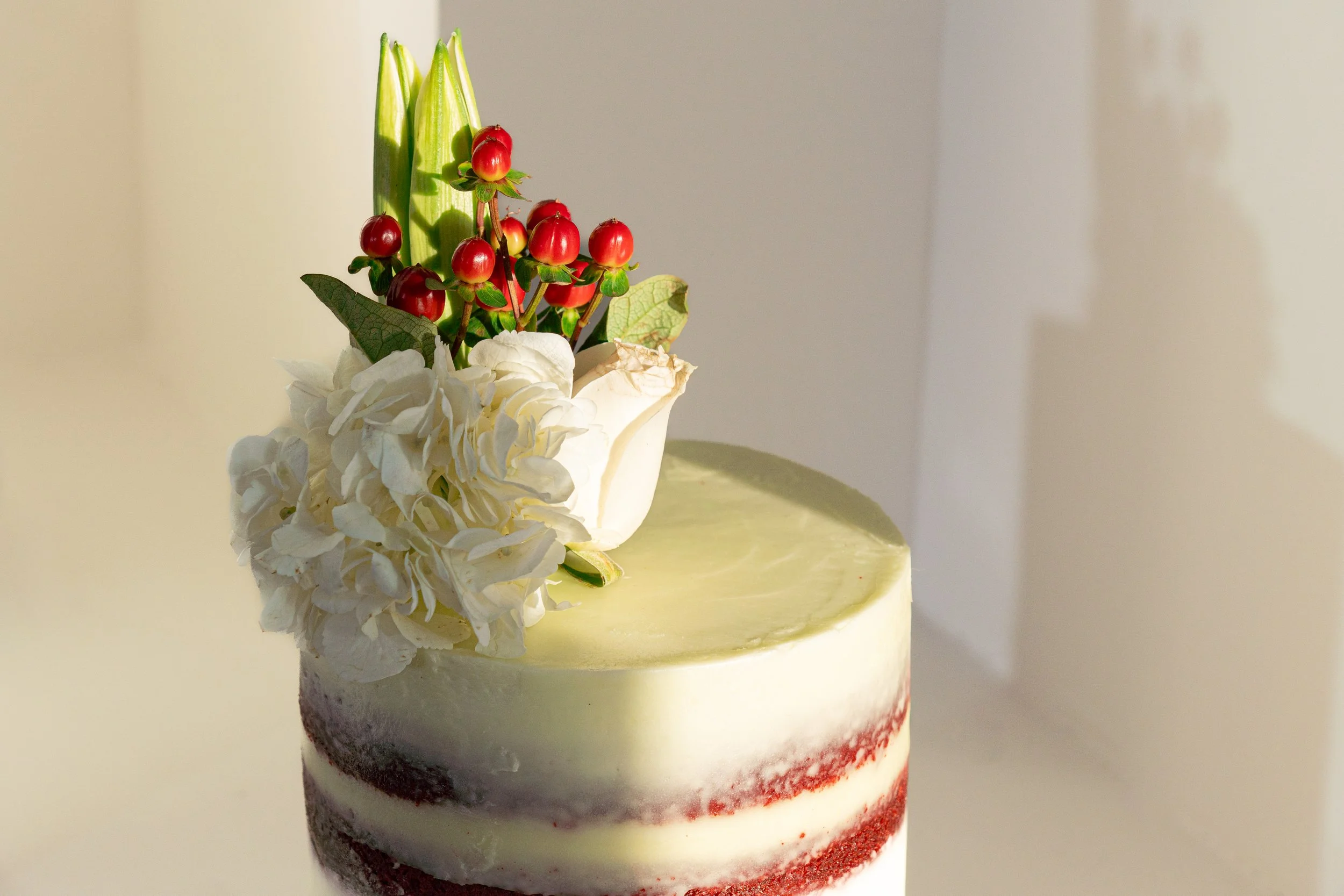 A layered cake decorated with white hydrangeas, green leaves, and clusters of red berries on top, with a smooth light-colored icing and a red velvet cake layer visible at the bottom.