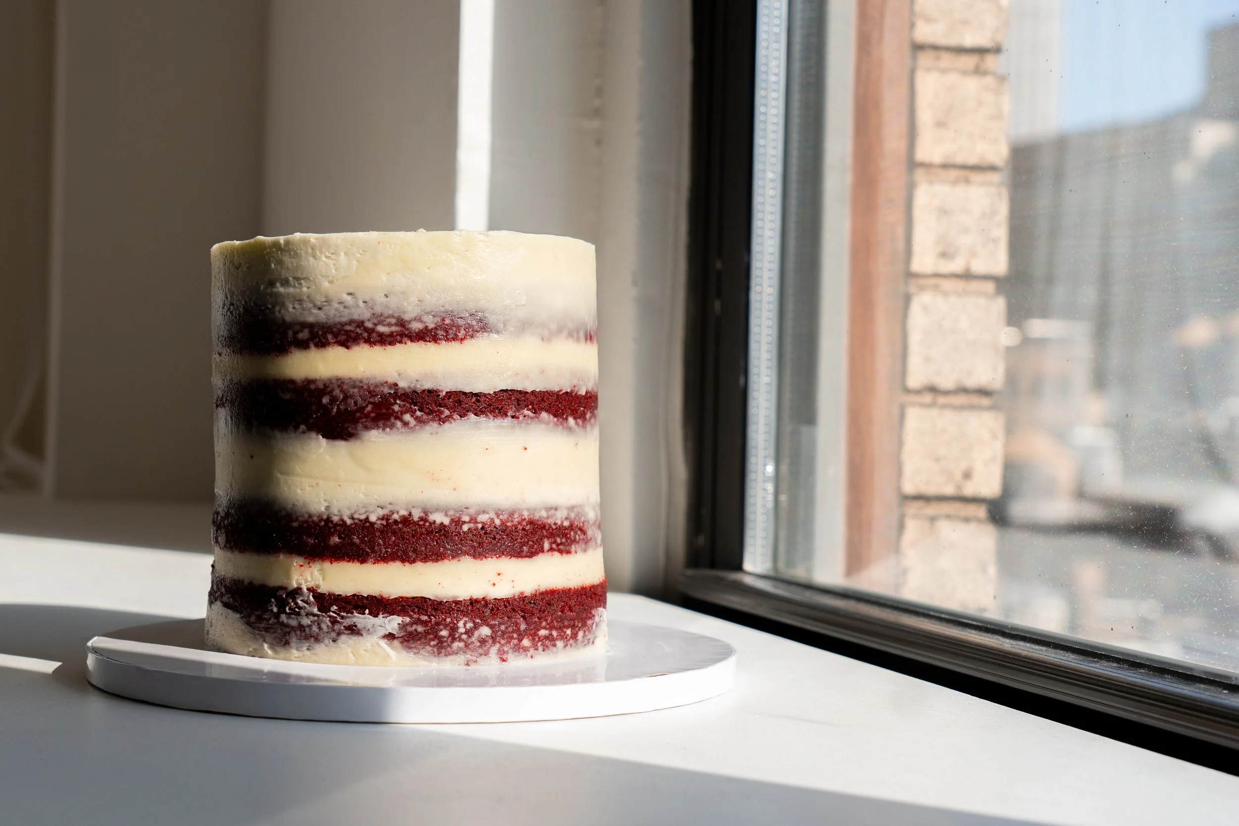 A layered red velvet cake with cream cheese frosting, sitting on a white cake stand near a window with sunlight.