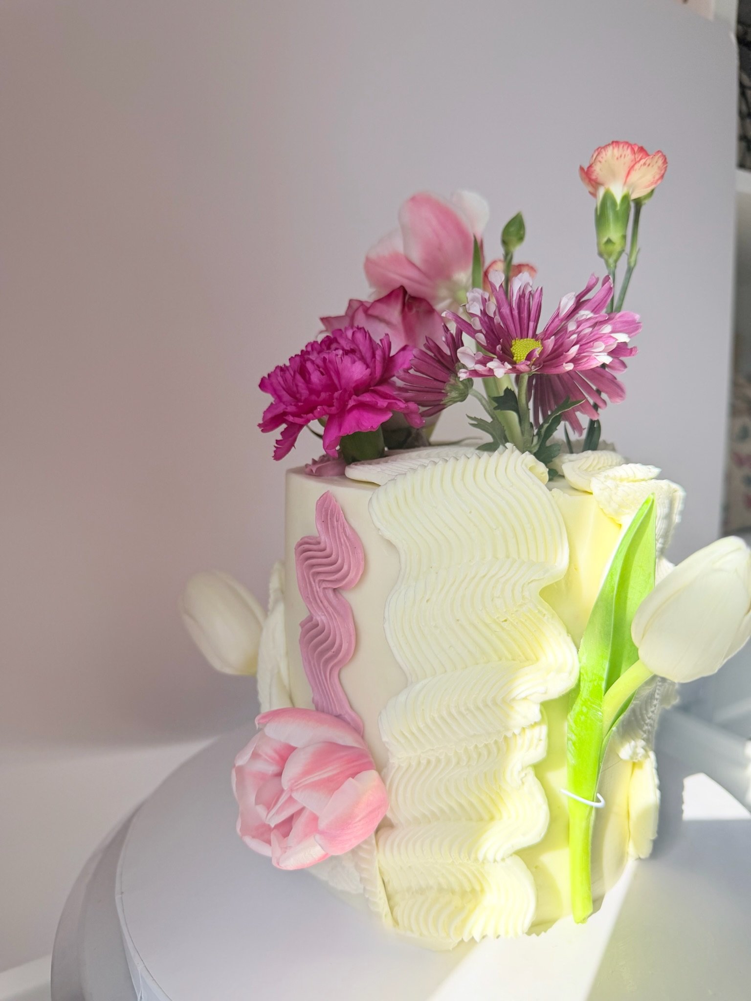 A floral cake with pink, purple, and white flowers decorated on top, placed on a white cake stand against a plain background.
