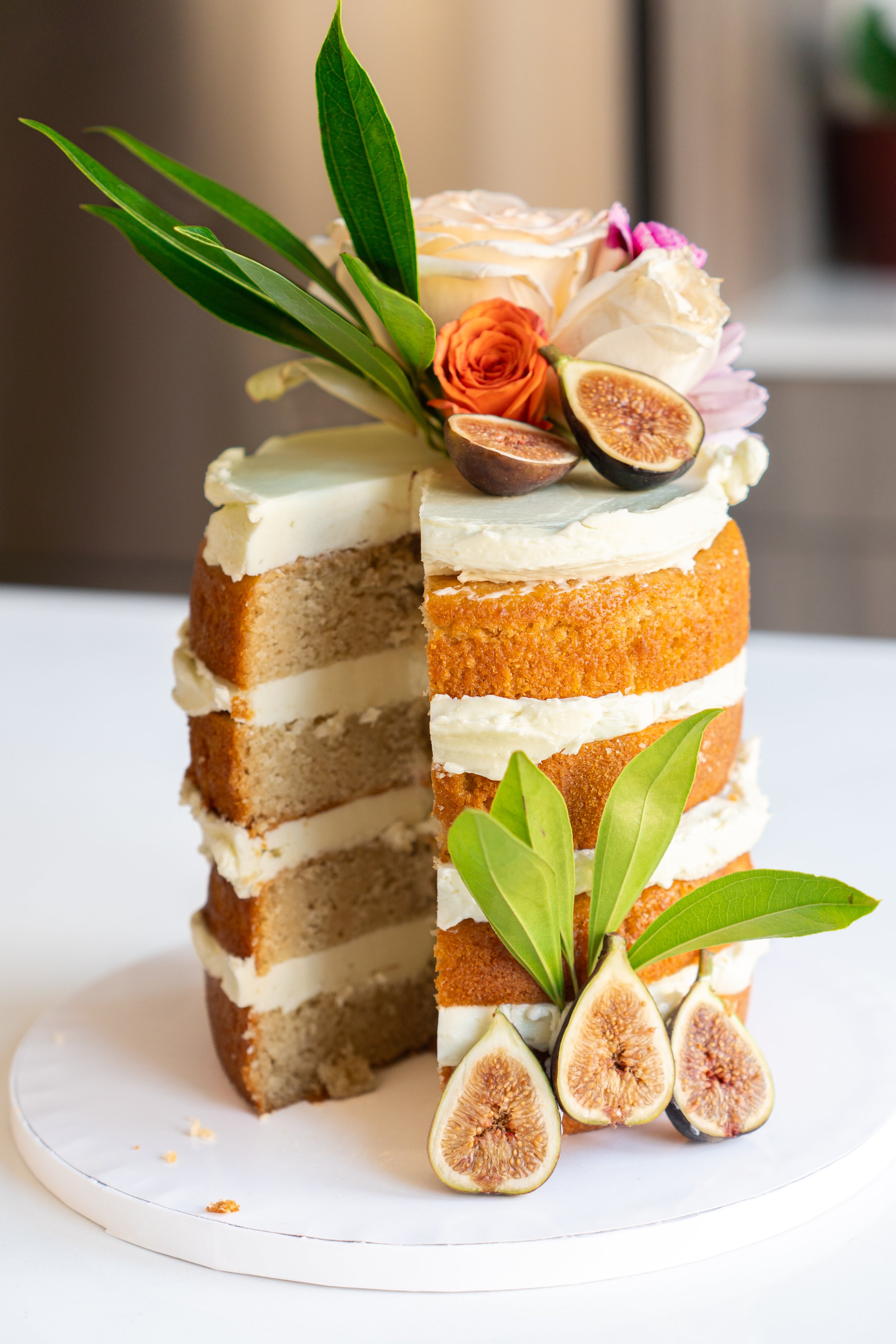 A multi-layered cake with white frosting, topped with fresh figs, orange roses, pink flowers, and green leaves, on a white cake stand.