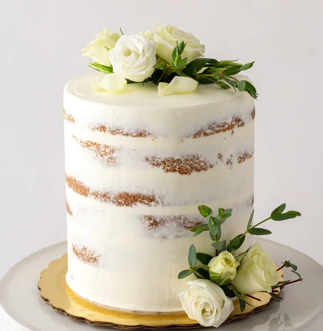 A white, semi-naked wedding cake decorated with white roses and green foliage, placed on a gold-colored base.