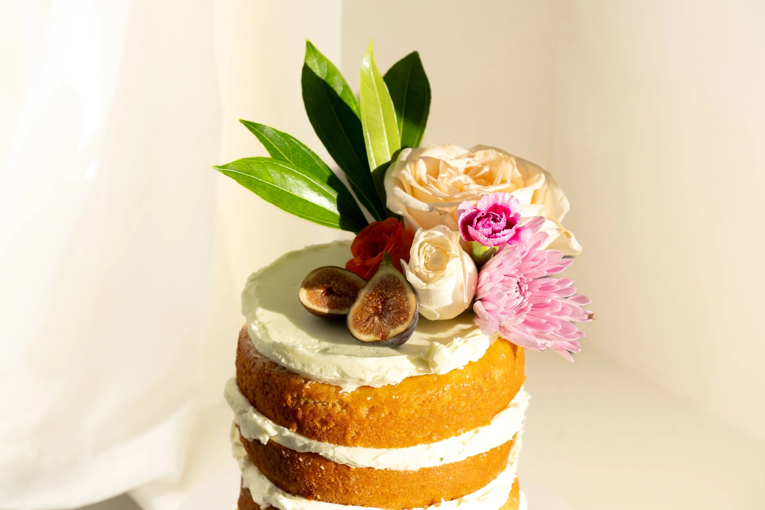 A layered cake with white frosting, topped with an assortment of flowers, figs, and green leaves.