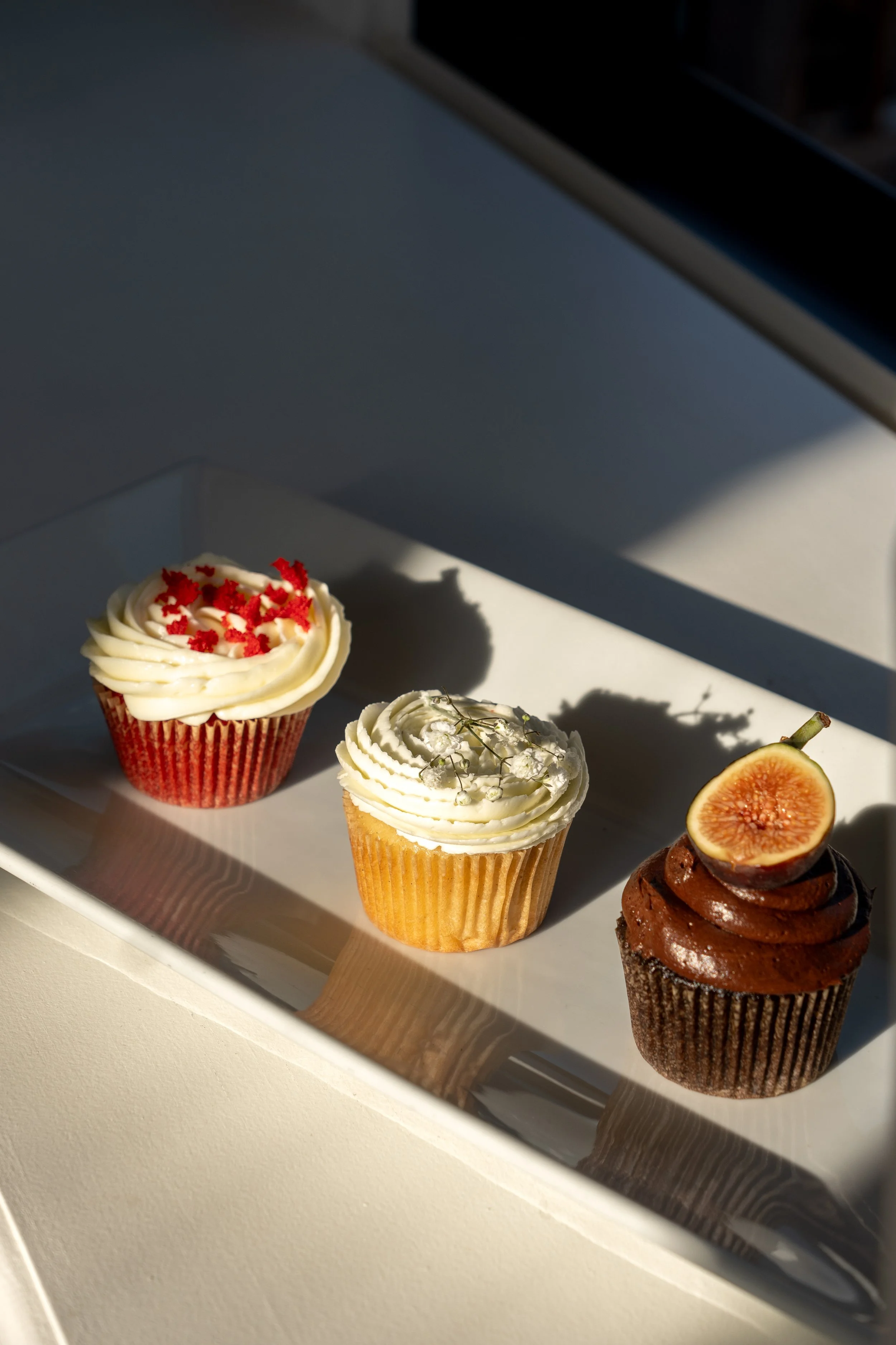 Assorted cupcakes with various toppings, including red sprinkles, white flowers, and a halved fig, displayed on a white tray.
