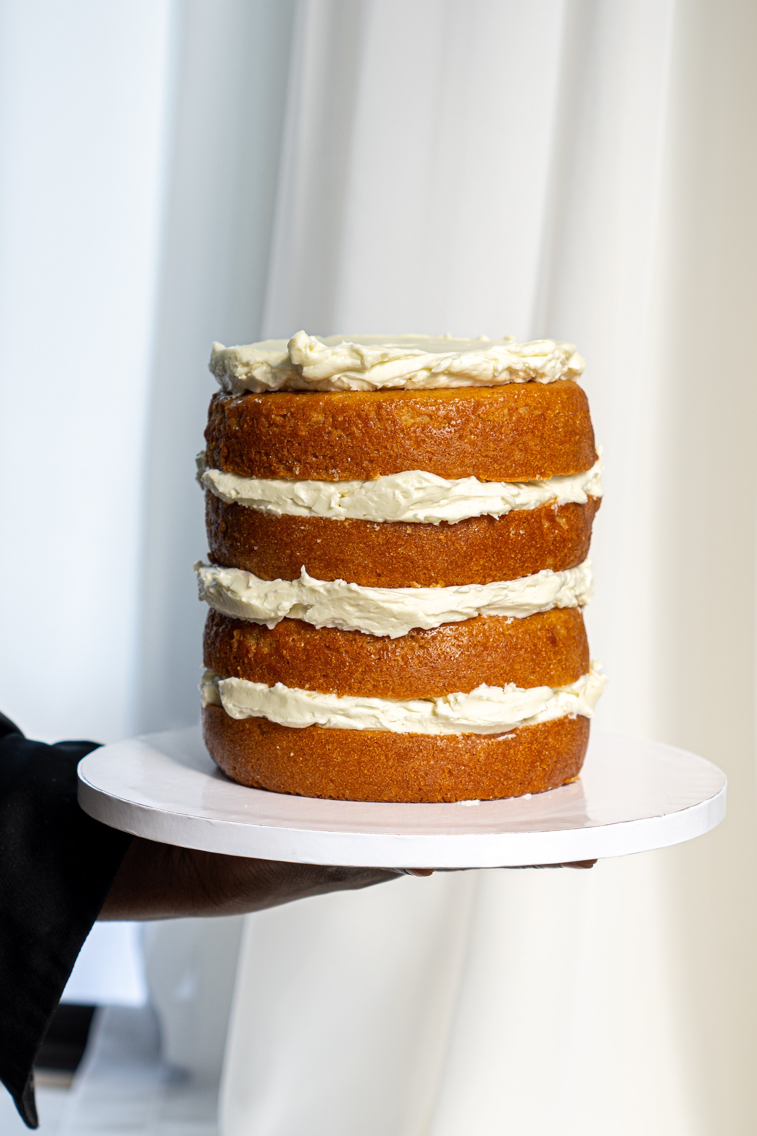 A person holding a multi-layered cake with white cream and caramel-colored layers, on a white cake stand.