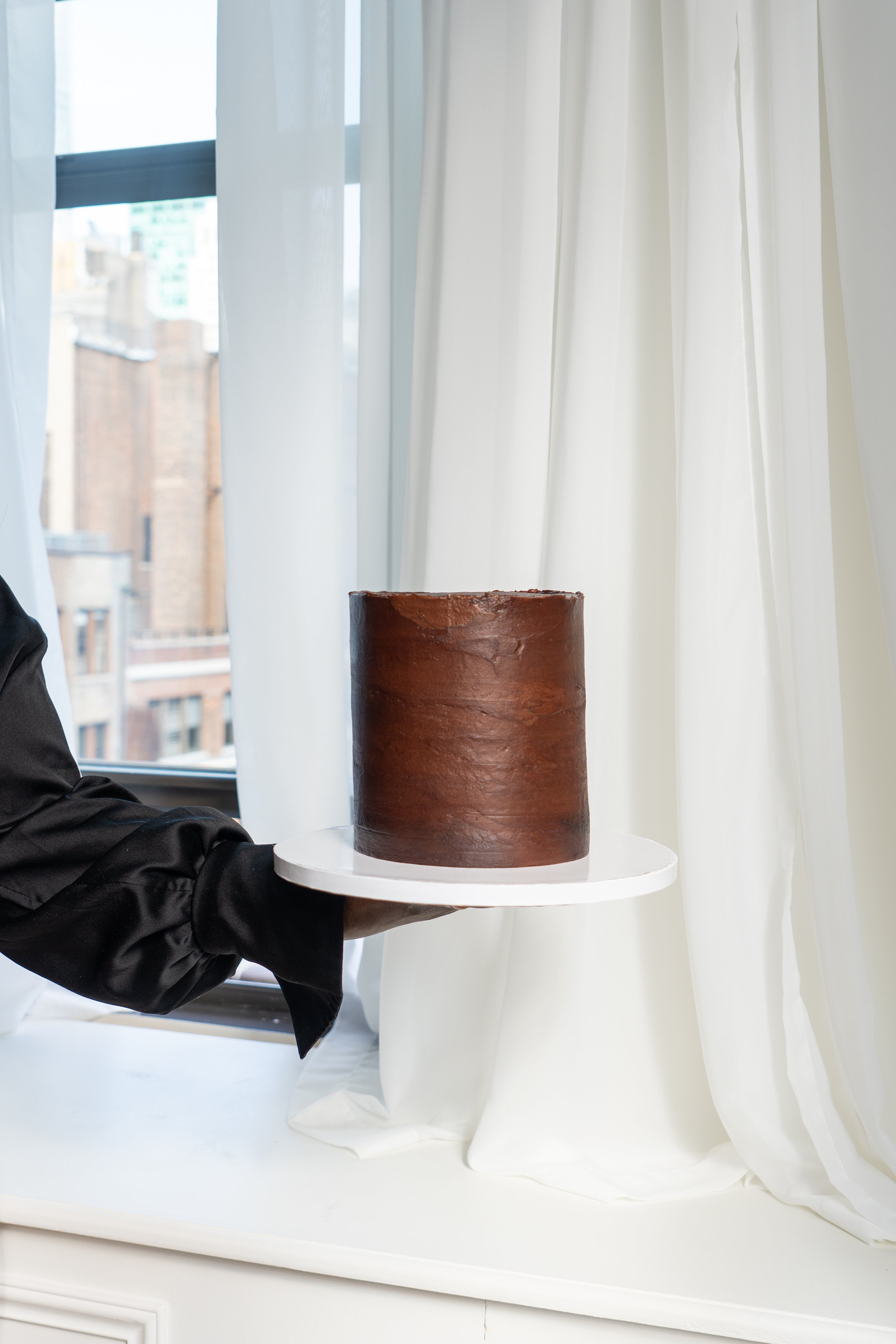 Person in black outfit holding a round wooden cake on a white serving board, standing near a window with white curtains, with a cityscape in the background.