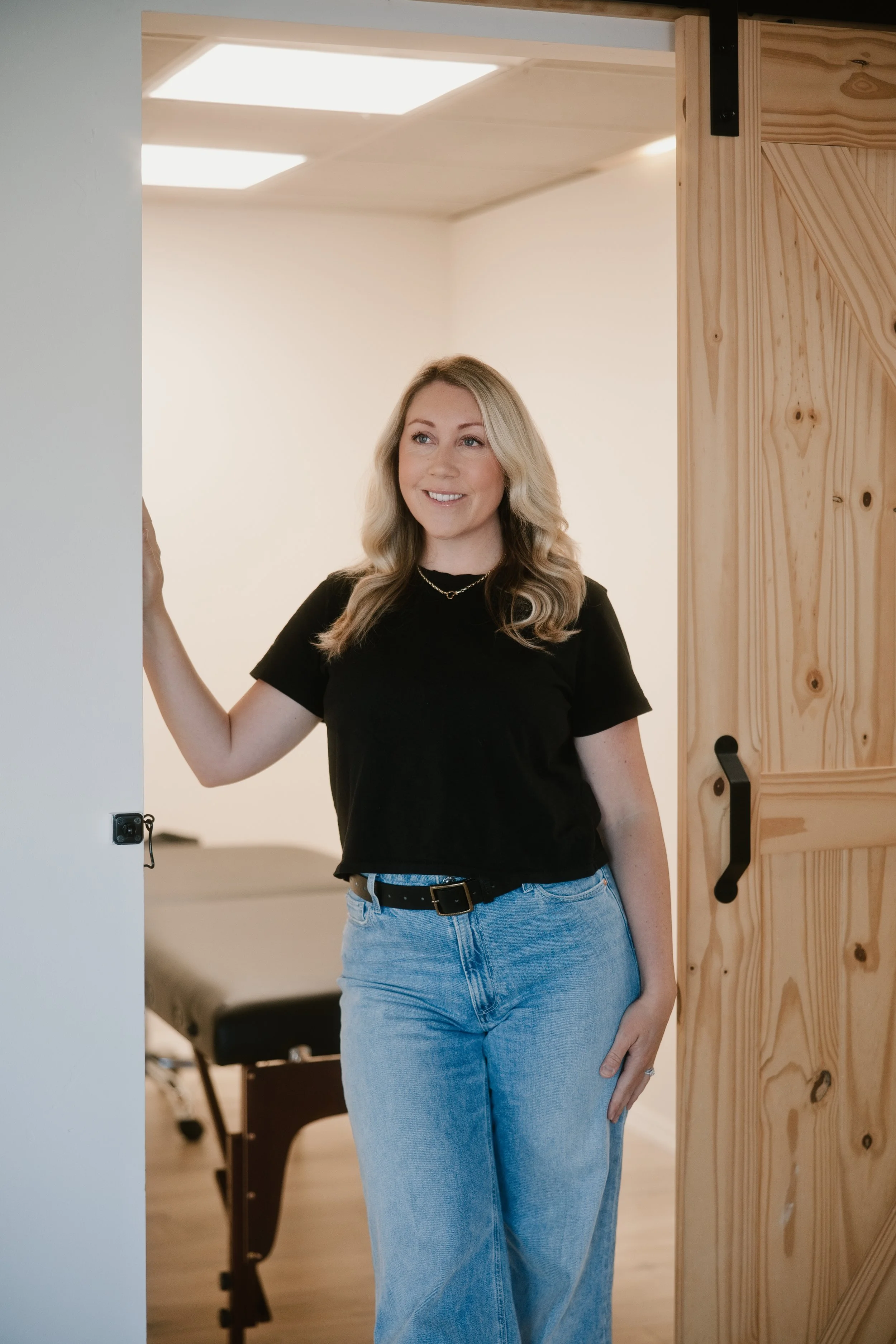 A young woman with blonde hair, wearing a black t-shirt and light blue jeans, standing in a doorway and smiling. The background shows a room with a chair and a dining table.