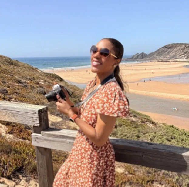Woman wearing sunglasses and a red floral dress smiling while holding a camera, standing on a wooden fence overlooking a sandy beach with cliffs in the background.