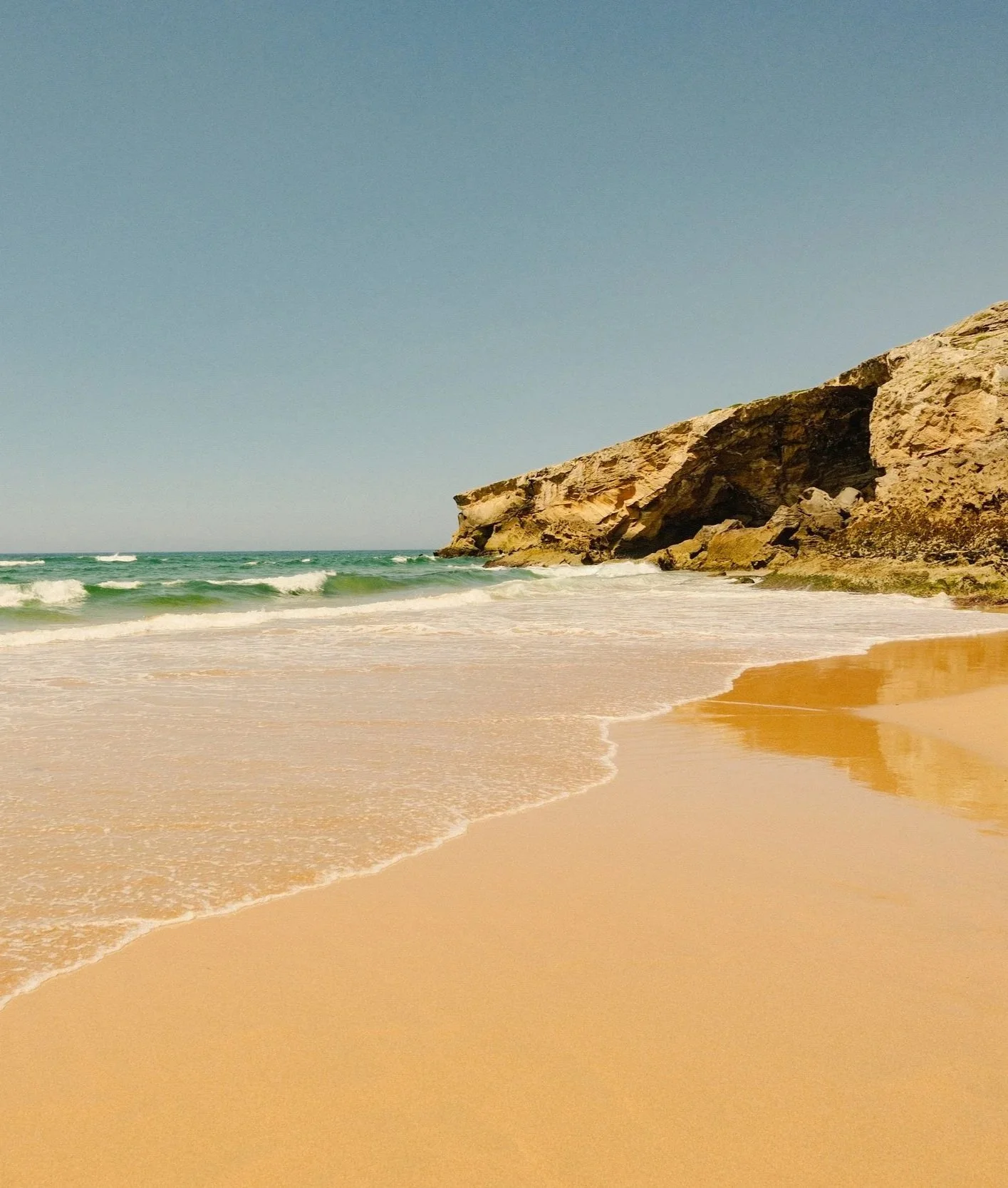 A sandy beach with gentle waves, a rocky cliff on the right, and a clear blue sky.