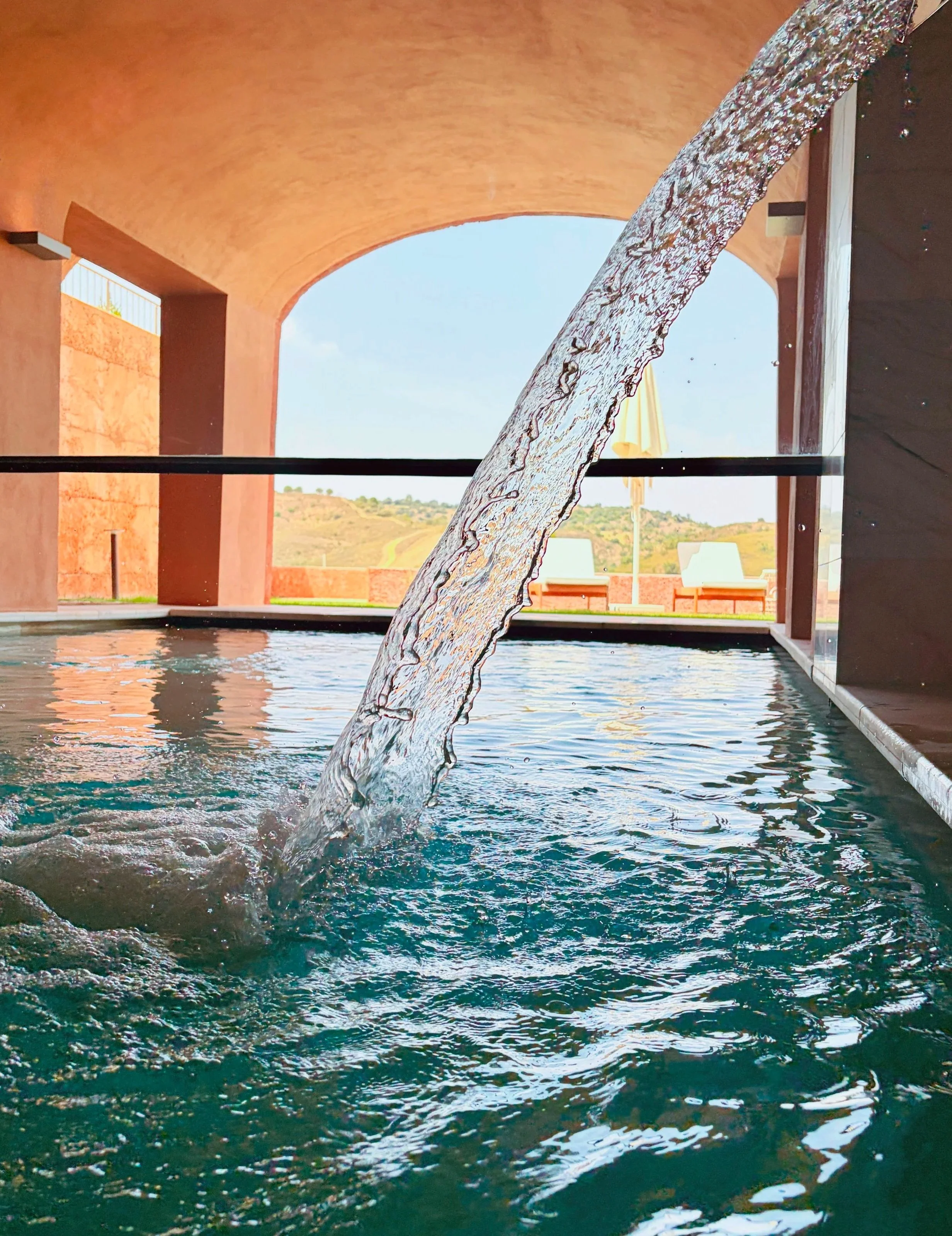 Indoor swimming pool with a waterfall feature, brown archways, and outdoor view with lounge chairs and an umbrella.
