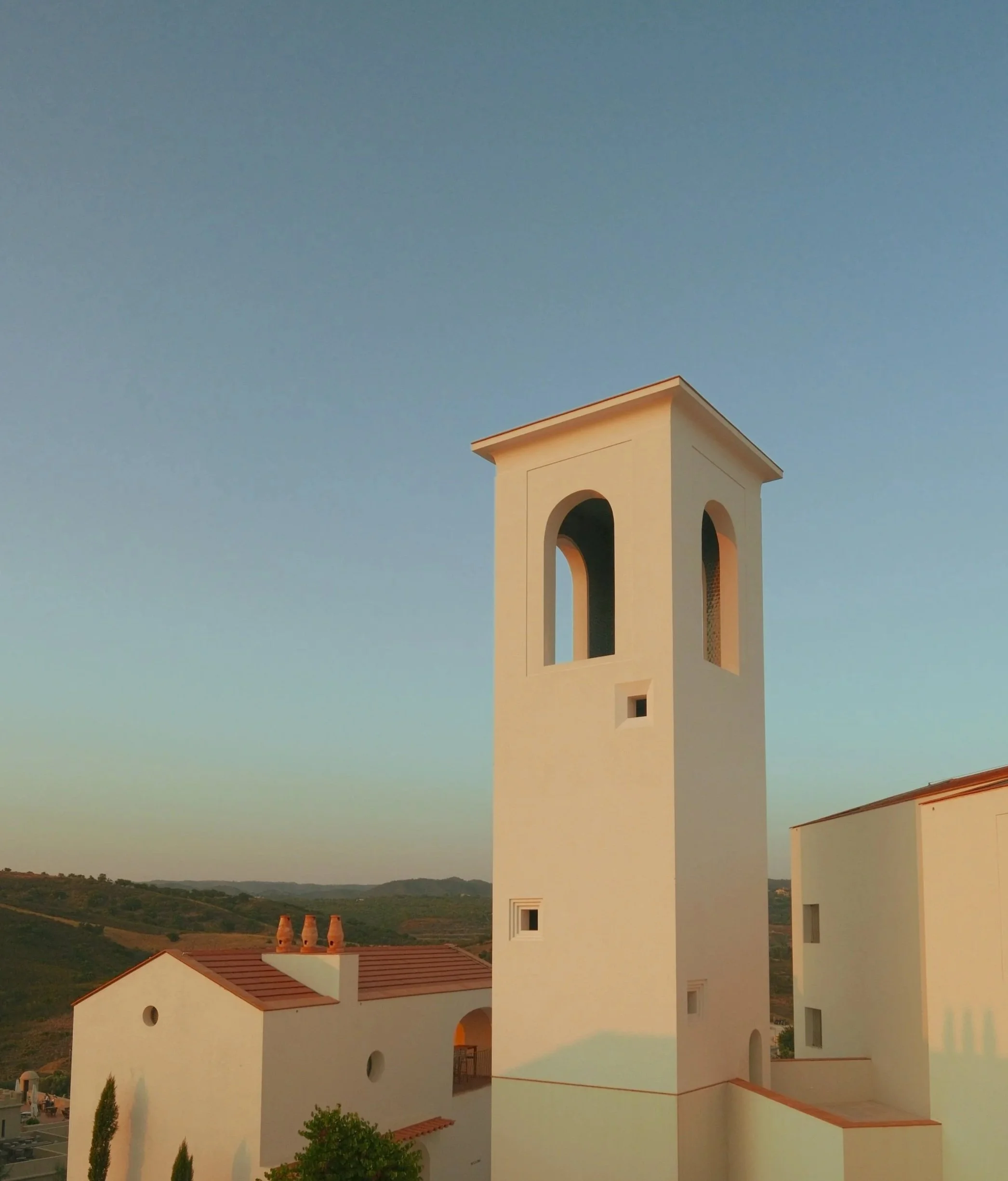 A white Mediterranean-style building with a tower against a clear blue sky, with hills in the background.