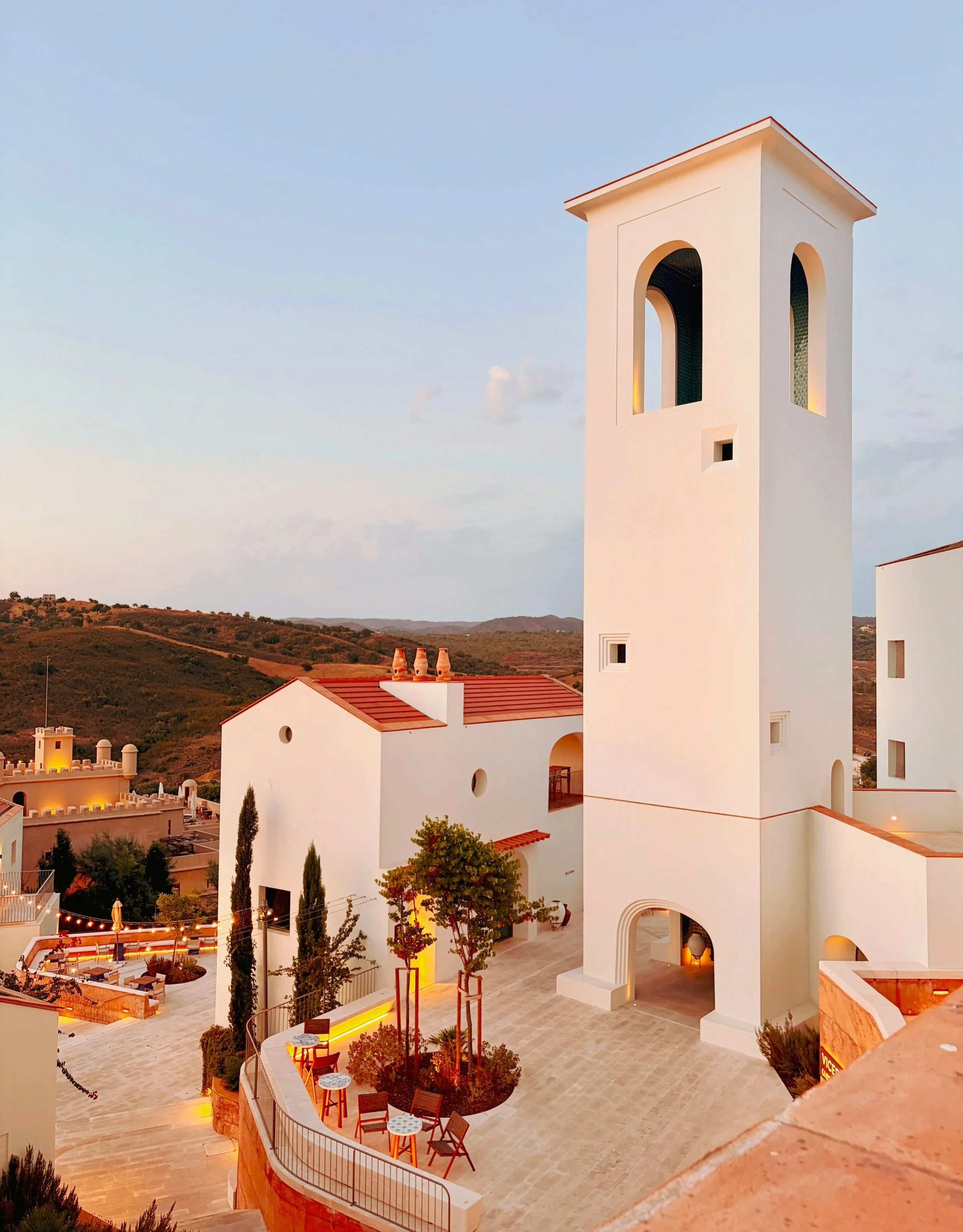 A view of Viceroy at Ombria Algarve with its Mediterranean-style buildings with a tall tower, surrounded by outdoor seating areas and greenery, at sunset.