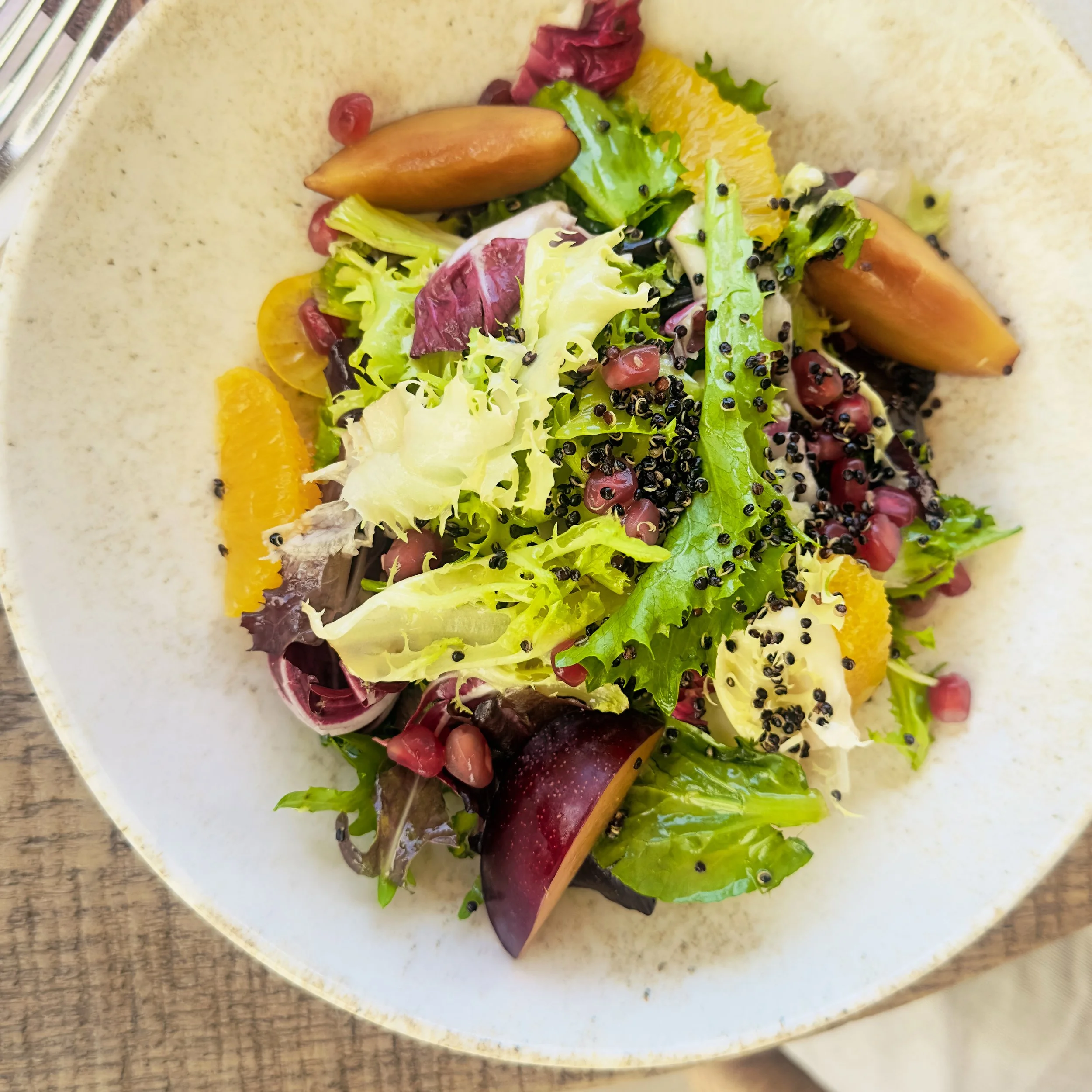A salad with a variety of greens, orange slices, pomegranate seeds, and black sesame seeds on a white plate.