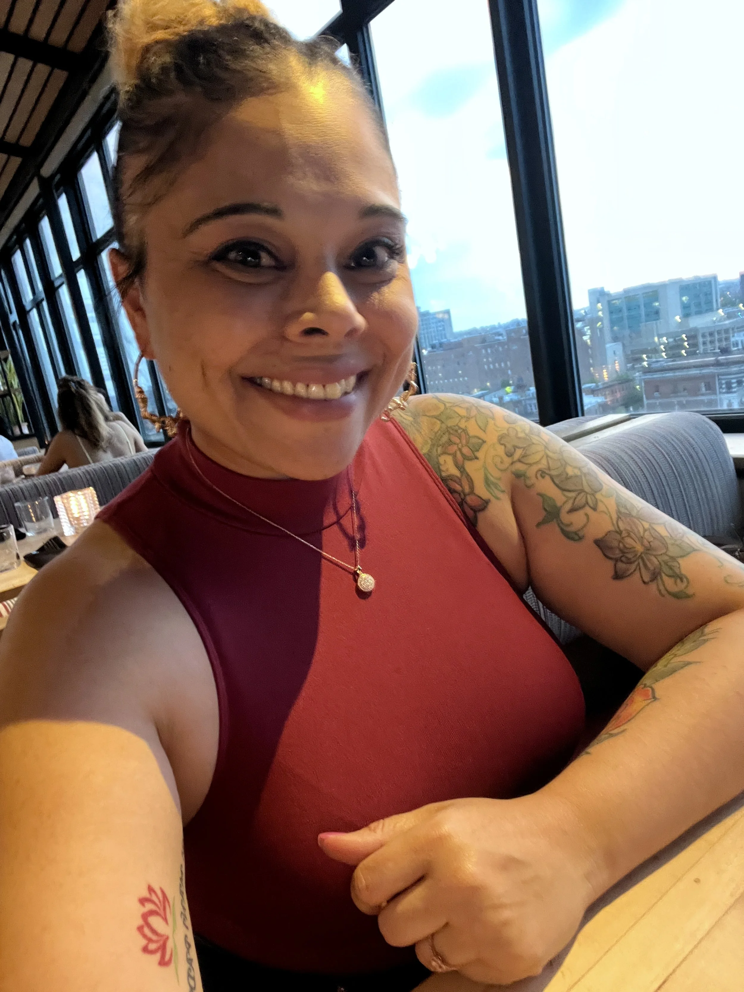 A woman with tattoos on her arm smiling and taking a selfie in a restaurant. She has a bun hairstyle, is wearing a red sleeveless top, and is in front of a large window overlooking a cityscape.