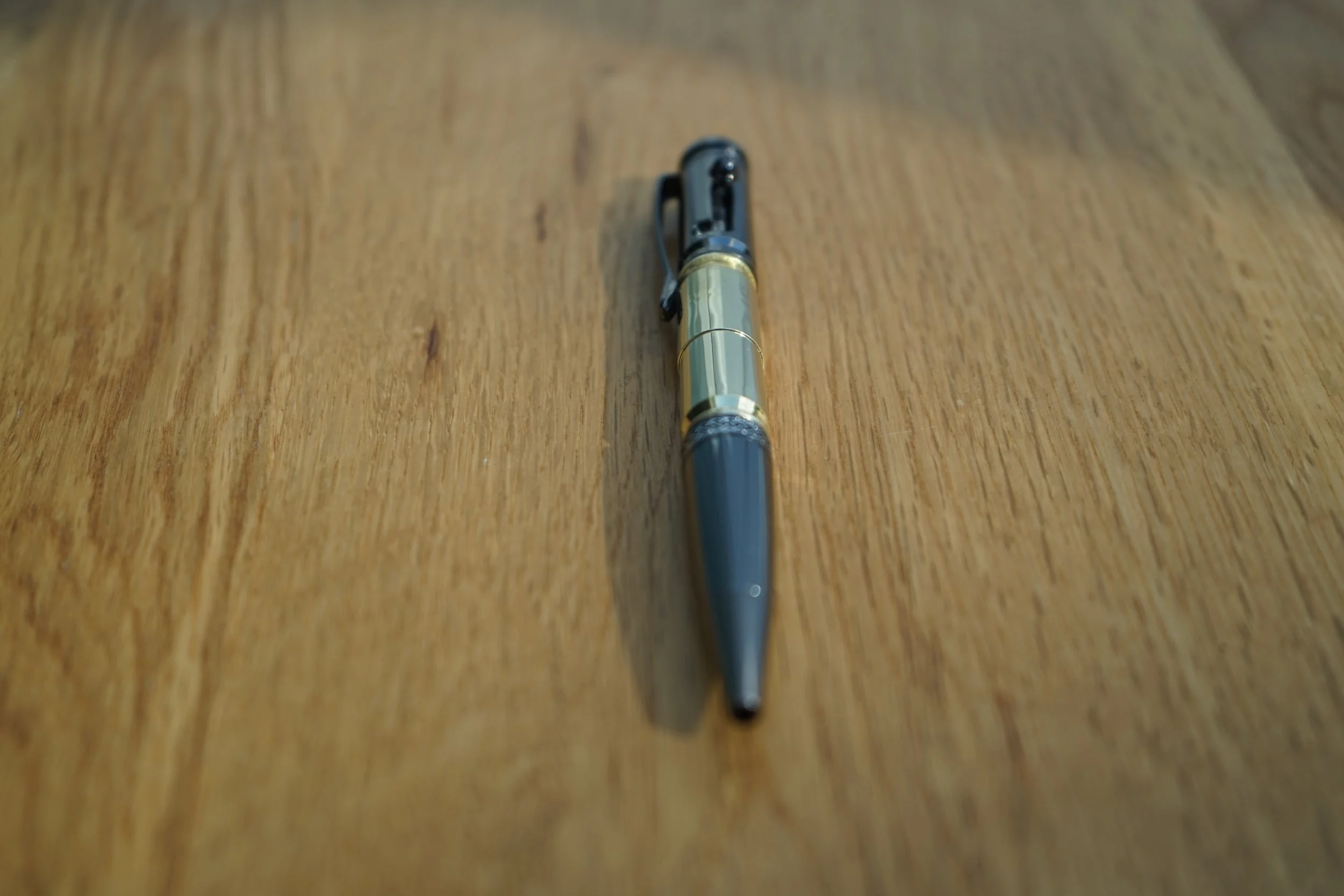A close-up of a bolt action ballpoint pen with a black clip, once fired brass and black body, lying on a wooden surface.