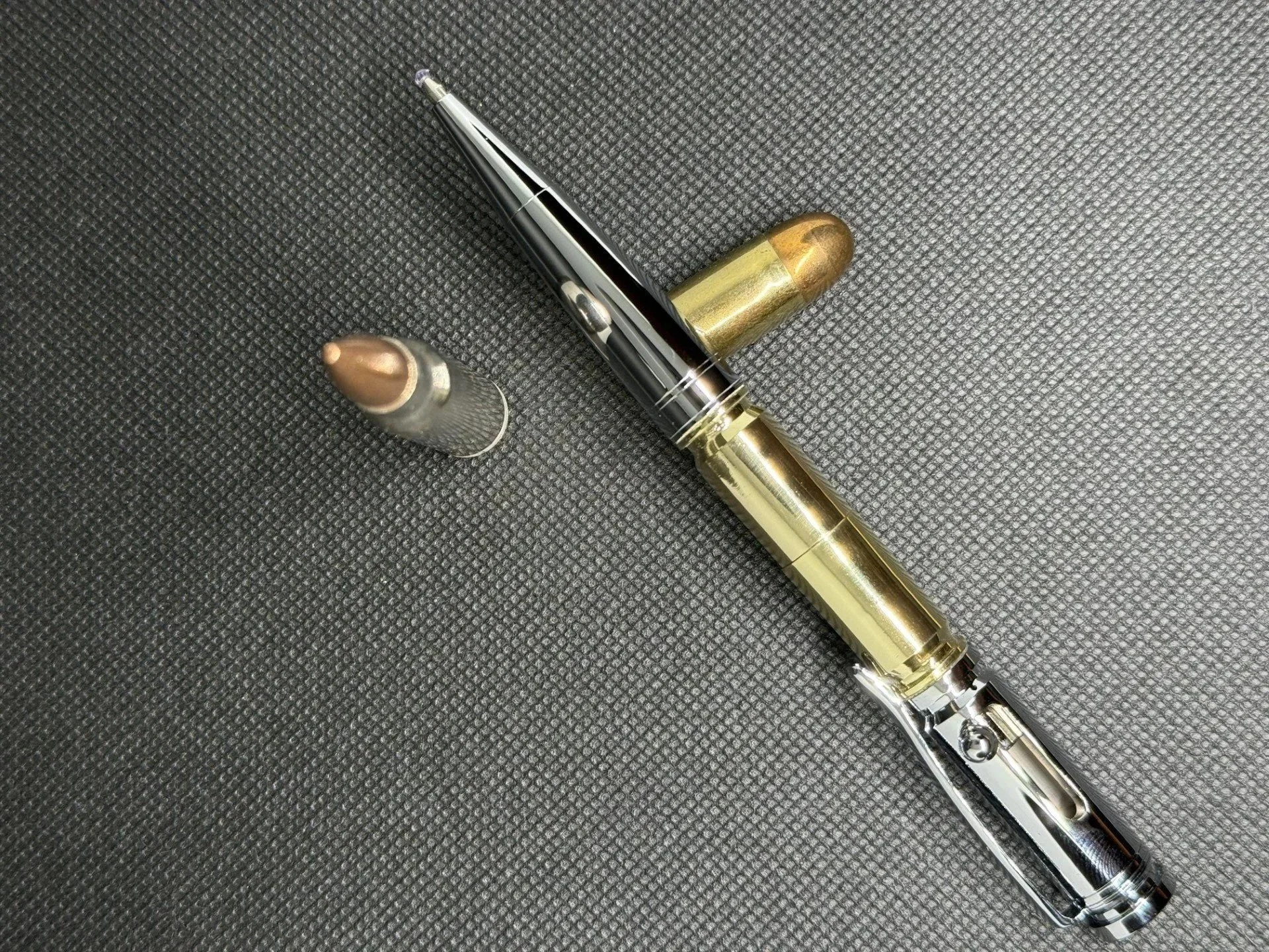 Close-up of a ballpoint bolt action pen with gold accents and two bullets resting on a textured black surface.