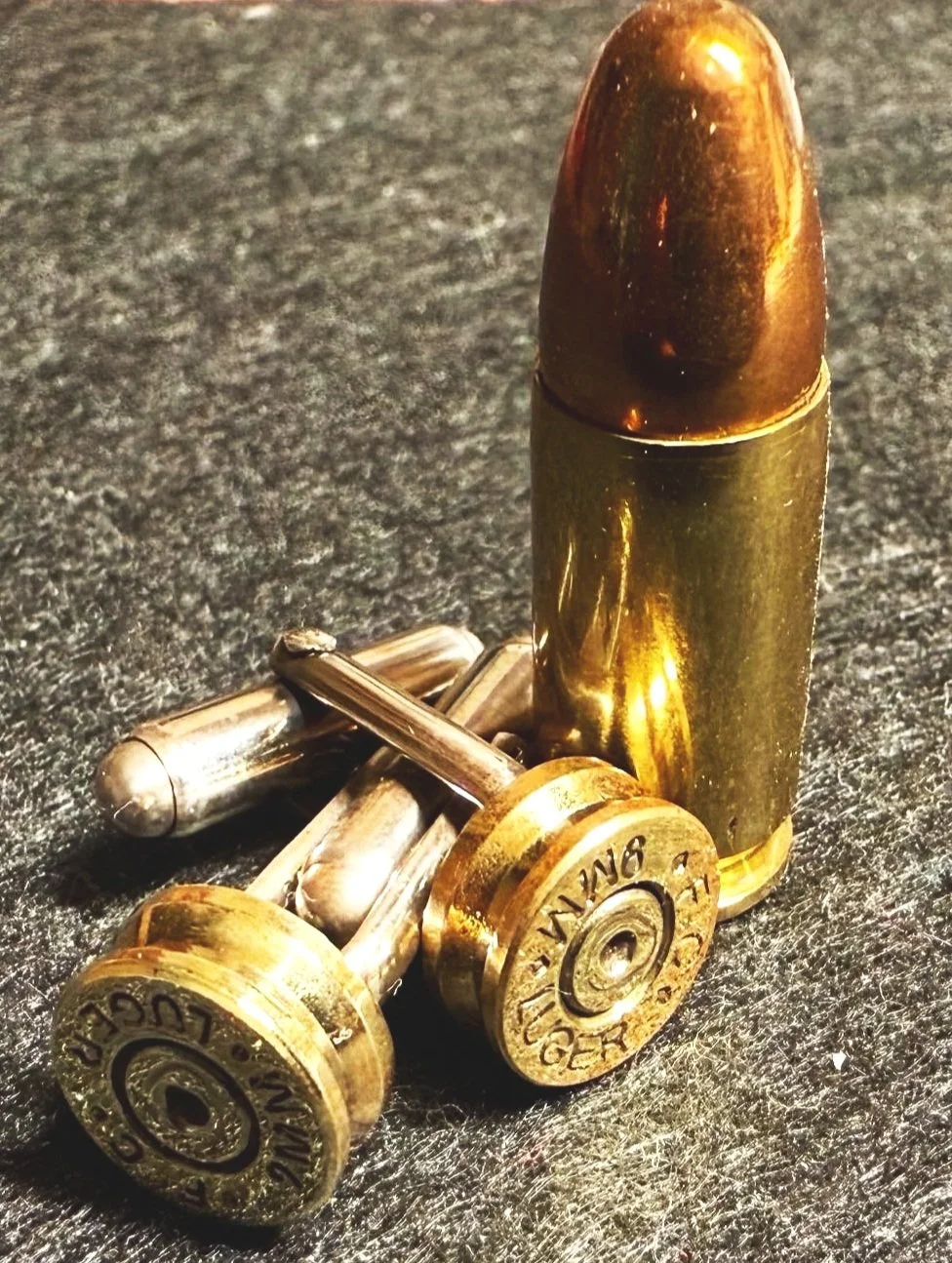 A bullet standing upright next to a pair of small brass cufflinks made from bullets on a dark surface.