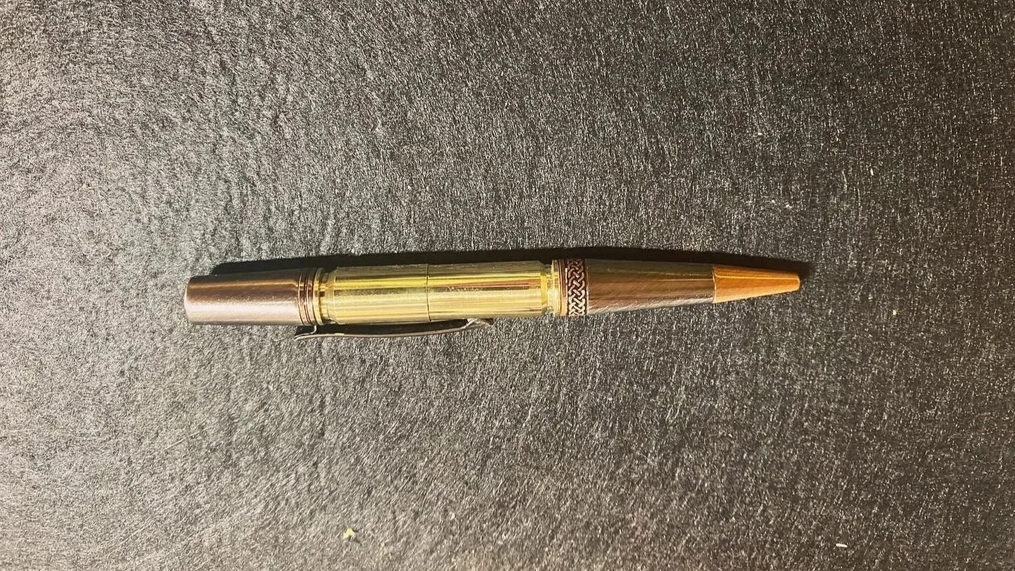 A pen with a metallic gun metal top, bullet casing middle, and gun metal tip, placed on a dark textured surface.