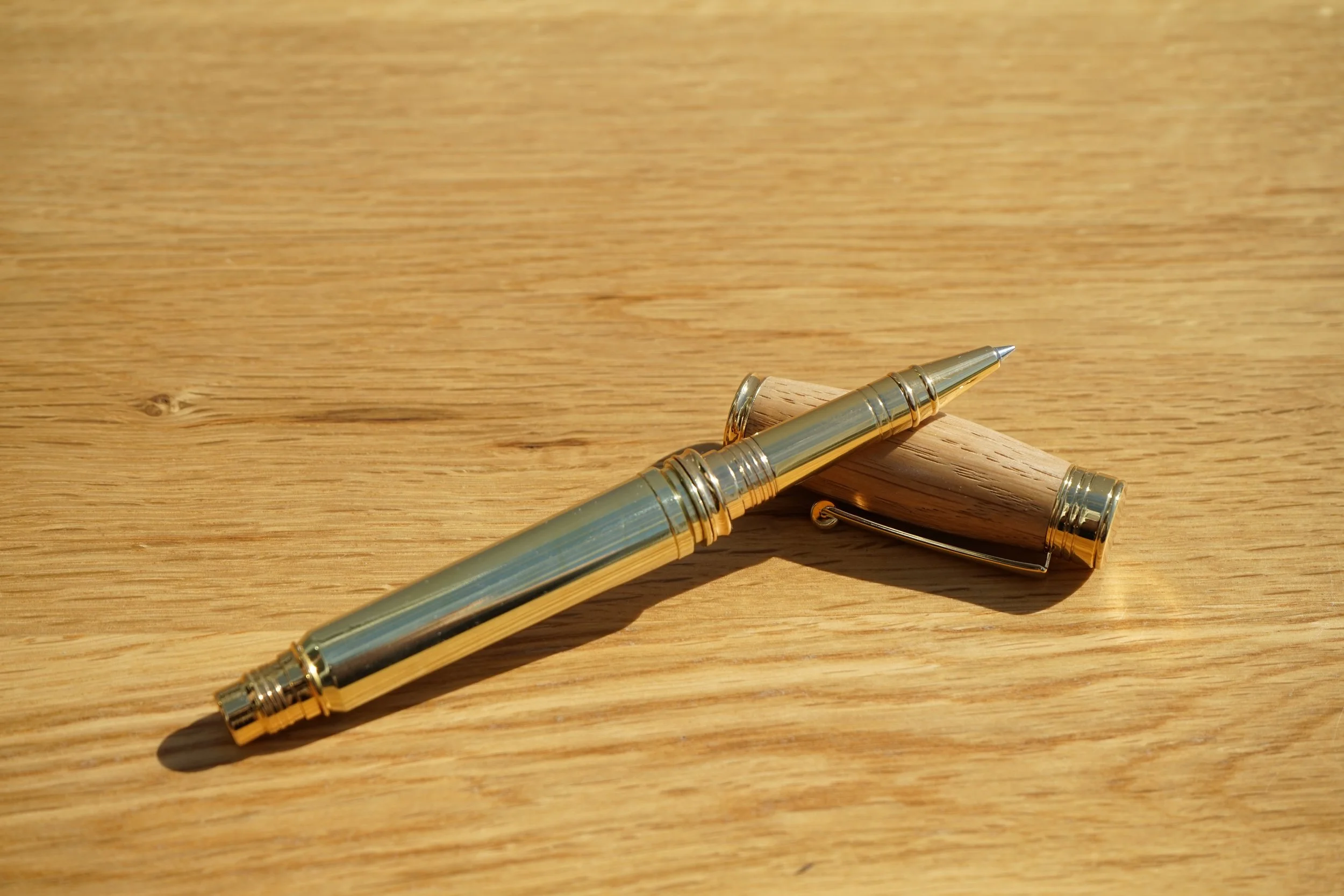 A gold fountain pen made with a once fired bullet casing and oak from a Dalmore Whisky cask on a wooden surface.