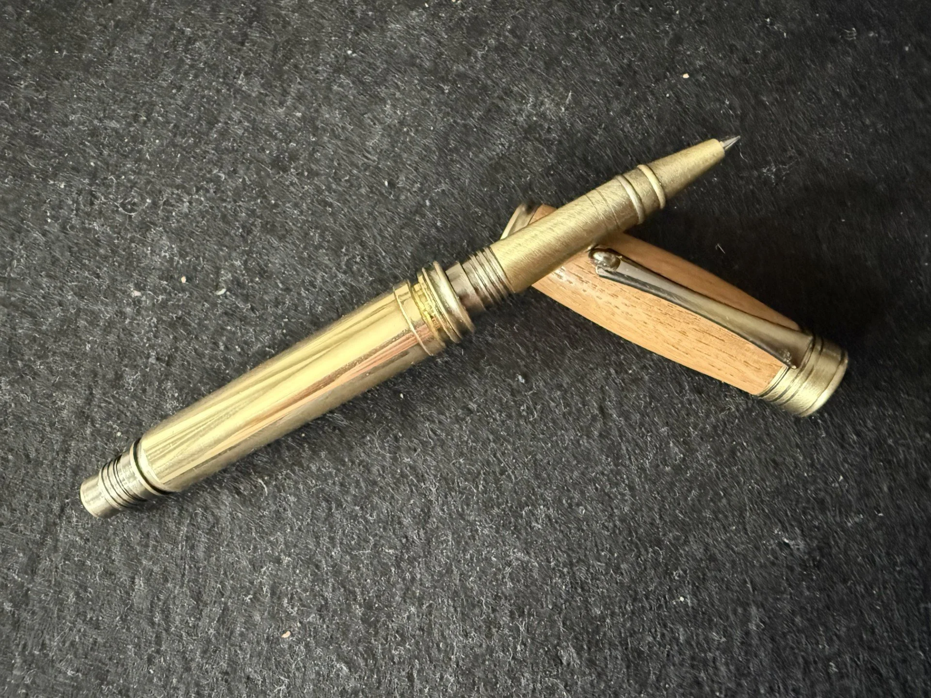 A brass or gold-colored fountain pen with intricate design details resting on a black textured surface, with a wooden pen lid partially visible underneath.