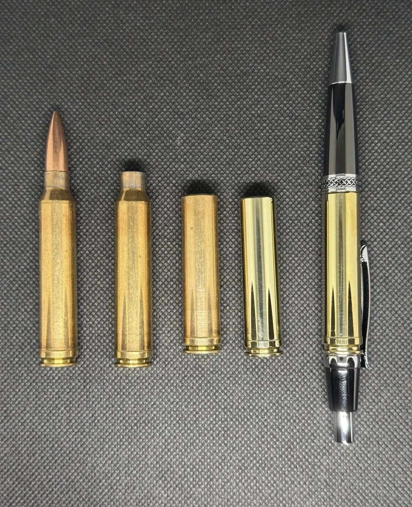 Four brass bullet casings and a bullet pen with a black and silver tip on a textured black surface.