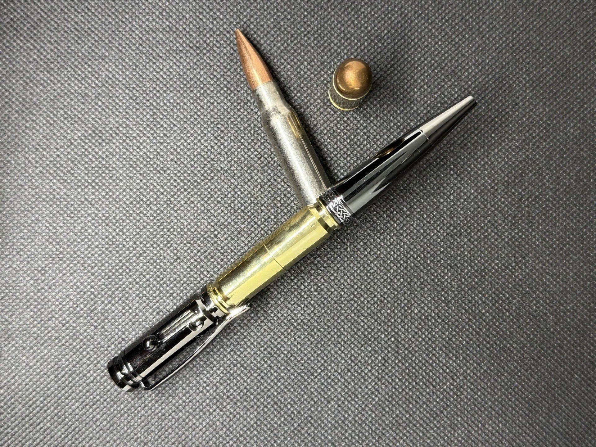 A silver and gold and gun metal bolt action bullet casing pen with two bullets on a dark surface.