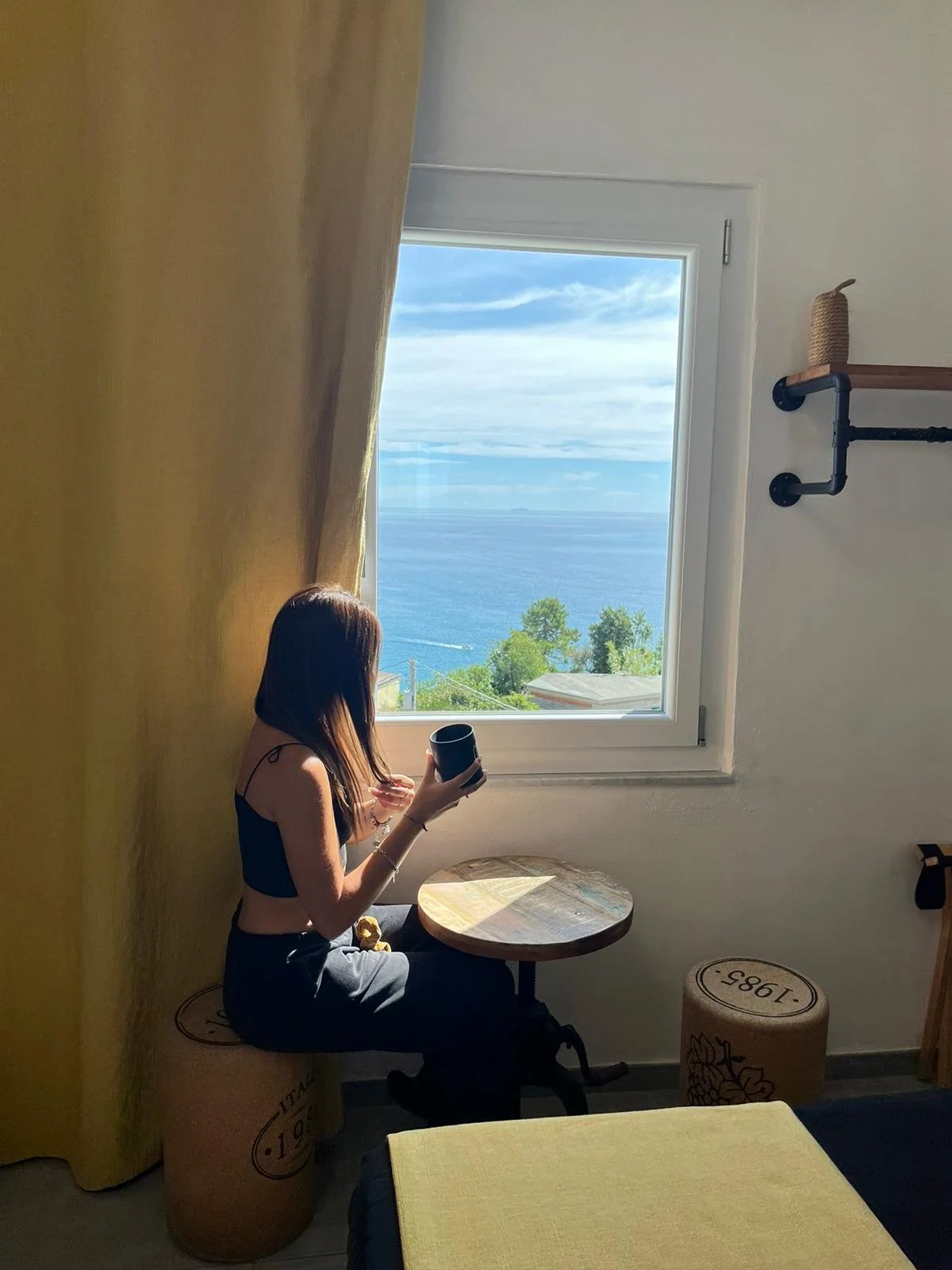 Una donna seduta vicino a una finestra che mostra una vista dell'oceano con cielo nuvoloso, ostacola parzialmente la vista con un tavolo tondo e un appoggio industriale.